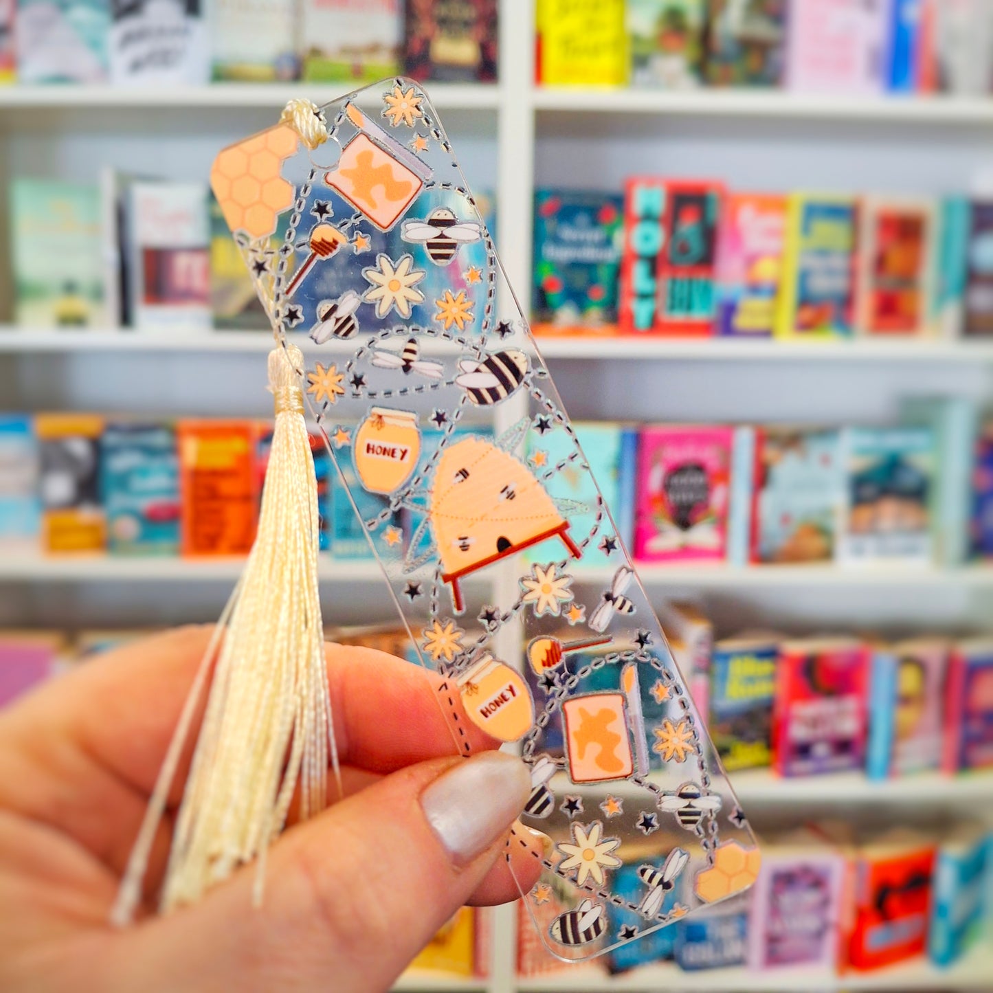 Transparent botanical bee bookmark with tassel held in front of a bookshelf.