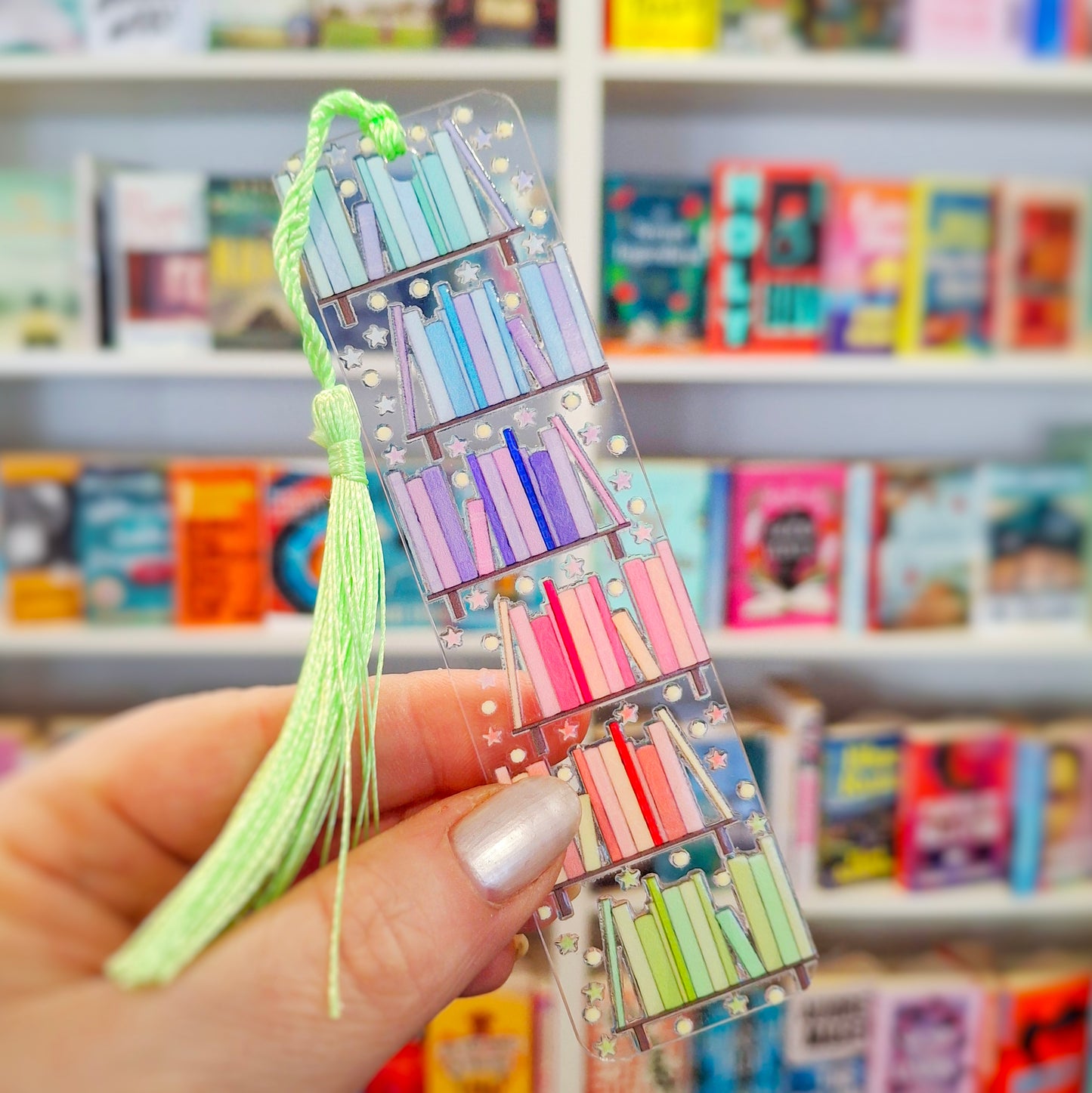 Transparent acrylic rainbow bookshelf bookmark with tassel held in front of a bookshelf.