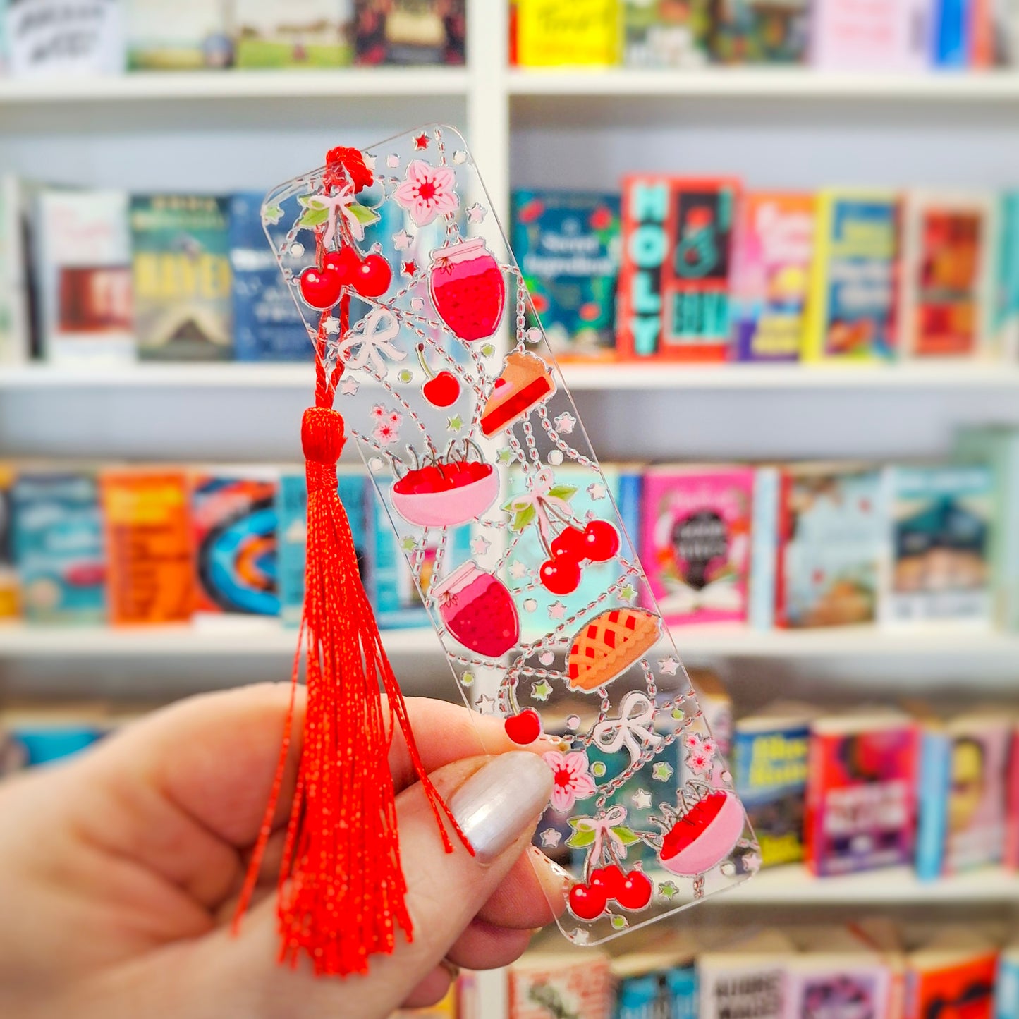 Transparent acrylic cute cherry pie bookmark with tassel held in front of a bookshelf.