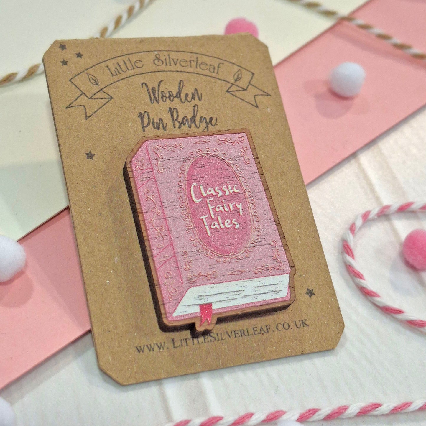 A wooden pin badge shaped like a pink classic fairy tale book with a pink ribbon bookmark, mounted on a brown kraft card that reads 'Little Silverleaf Wooden Pin Badge'. The badge is styled on a pink and cream background with decorative pom-poms and twine.