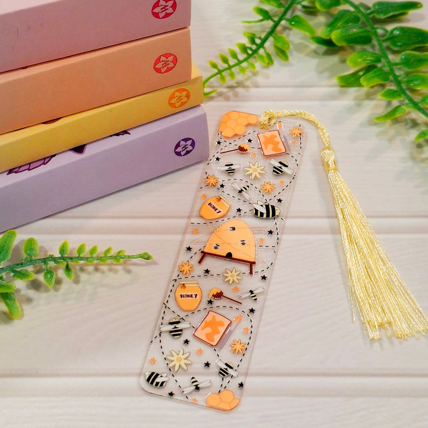 Honey bee acrylic bookmark with tassel on a light surface, with books and green leaves.