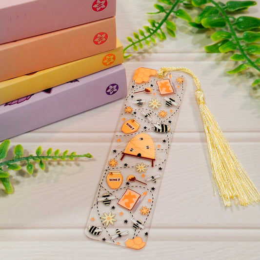 Honey bee acrylic bookmark with tassel on a light surface, with books and green leaves.