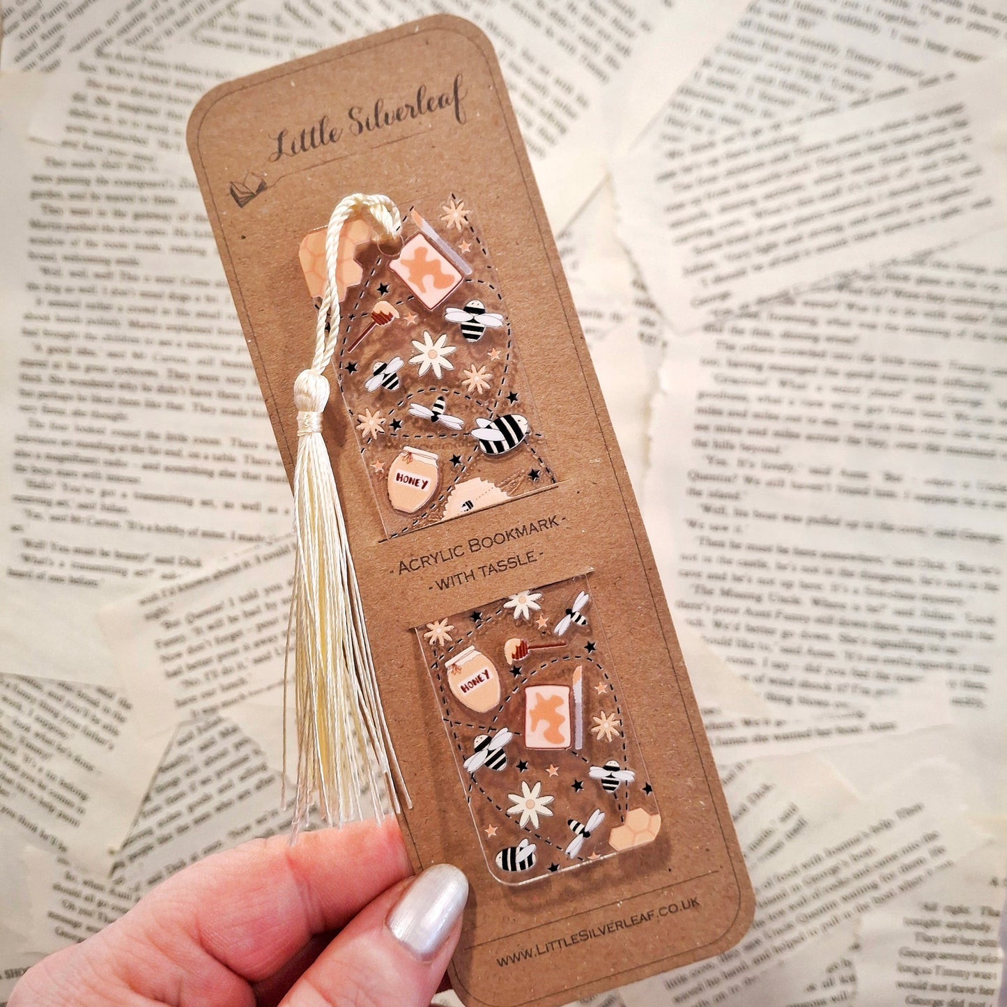 Acrylic bookmark with honey bee theme and a tassel, packaged on a brown kraft backing card, held over a book page background.