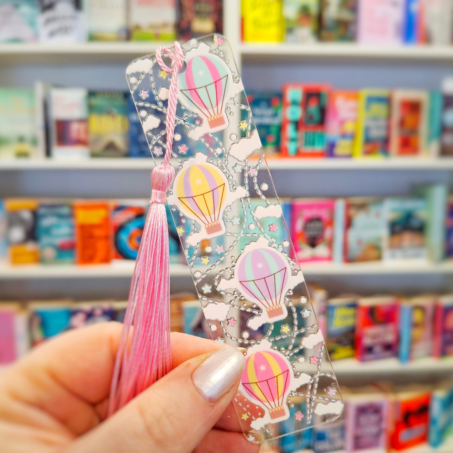 Transparent acrylic hot air balloon bookmark with tassel held in front of bookshelves.