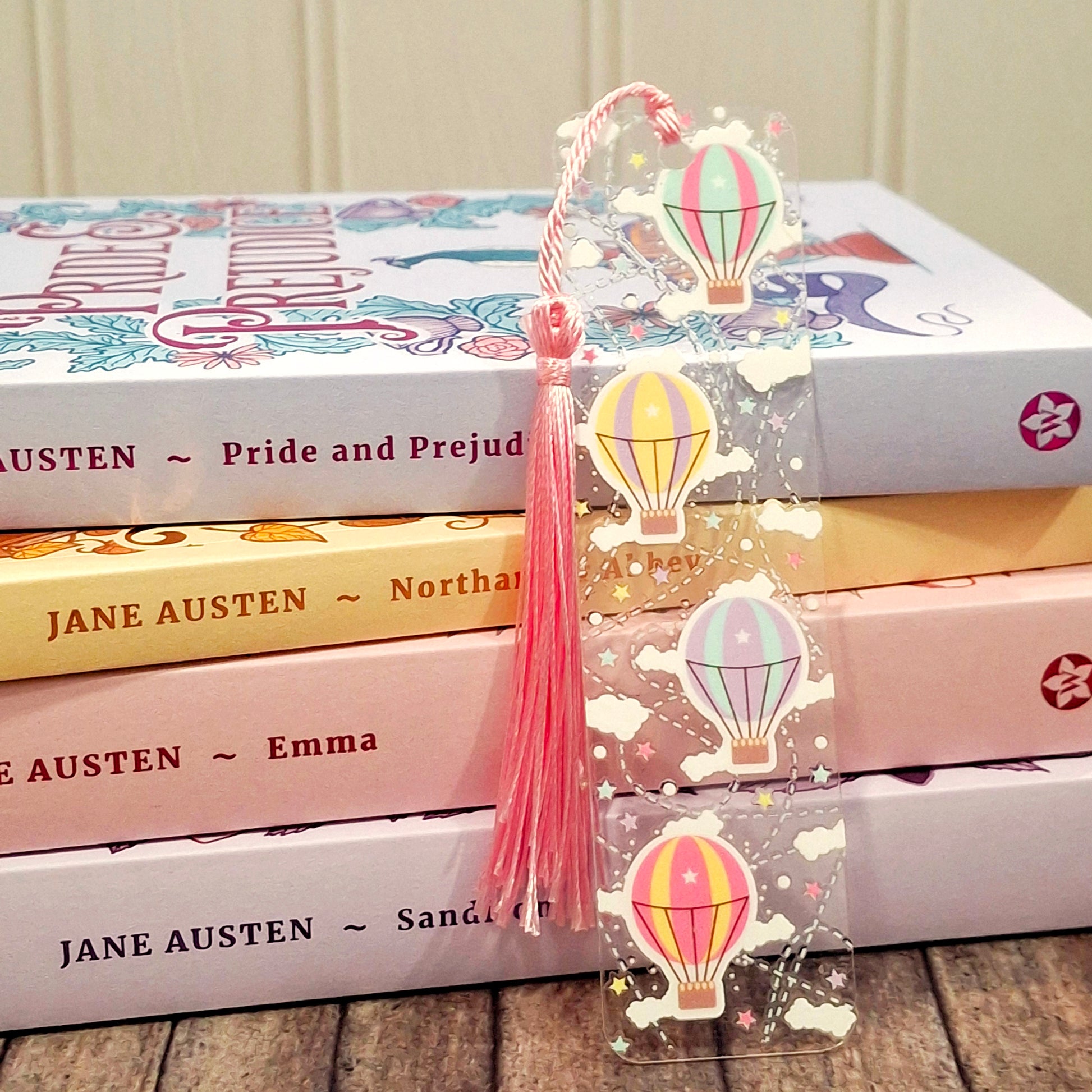 Travel inspired acrylic bookmark with hot air balloon illustrations and a tassel resting against a stack of books.