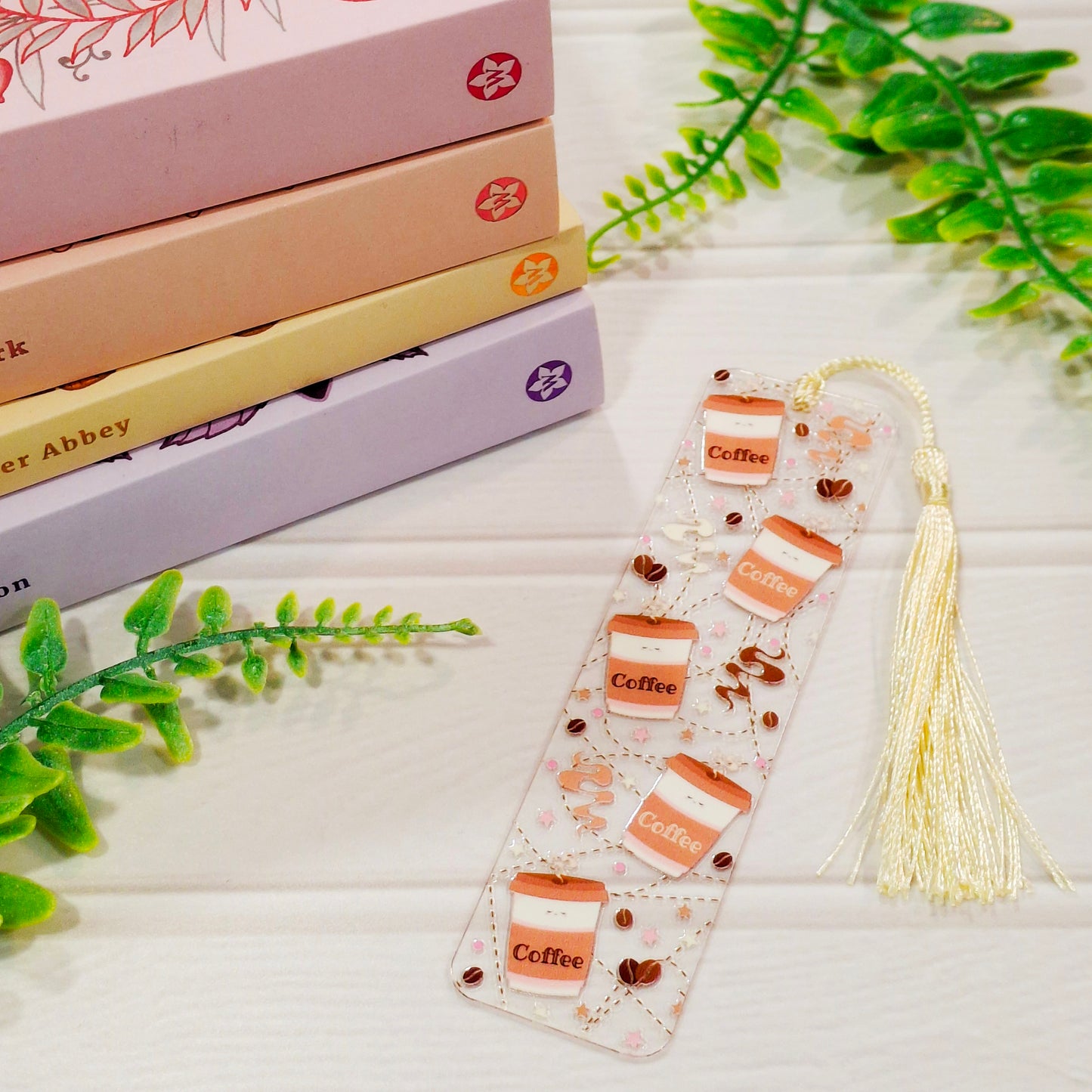 Coffee themed acrylic bookmark with tassel on a light surface, with books and leaves.