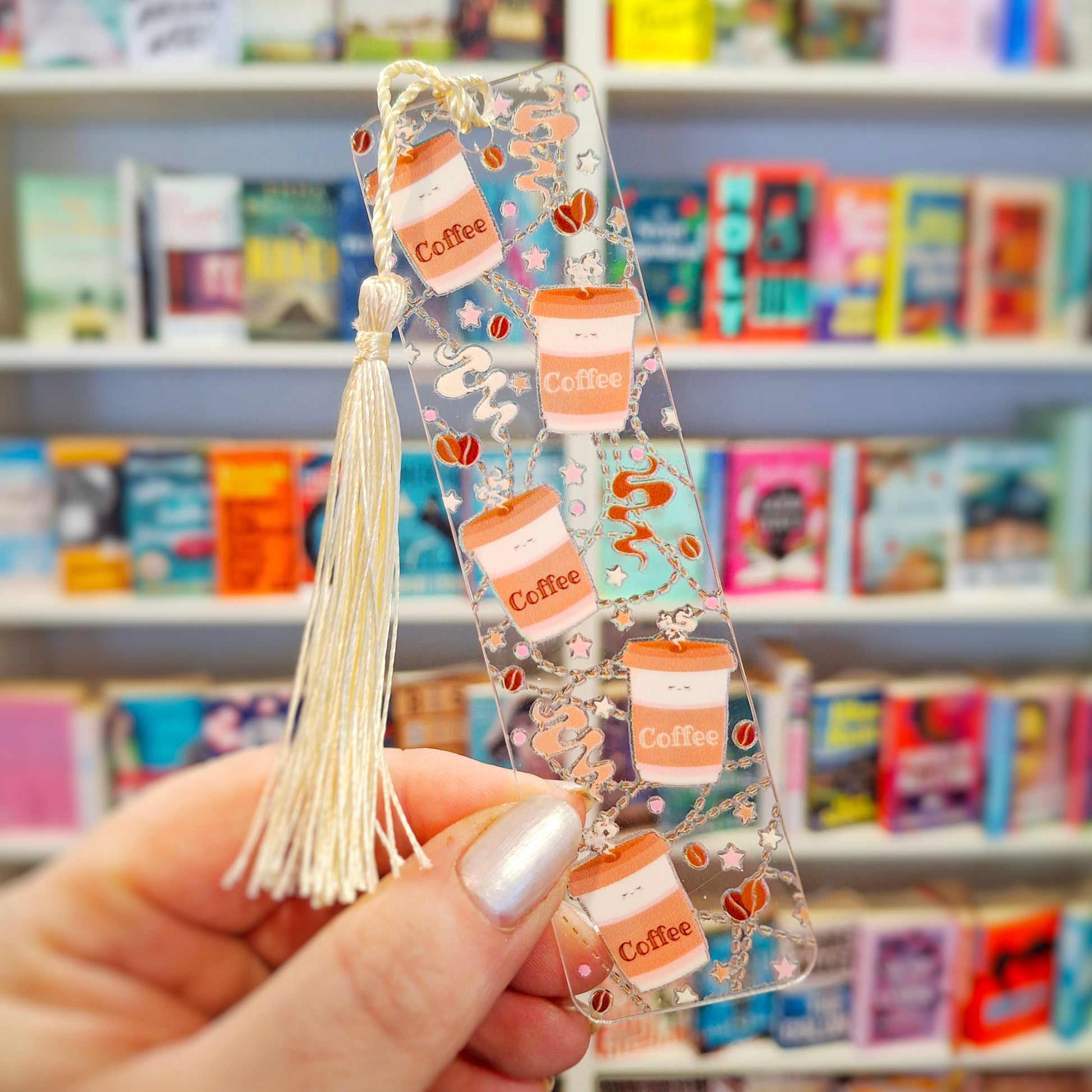 Transparent acrylic morning coffee bookmark with tassel held in front of a bookshelf.