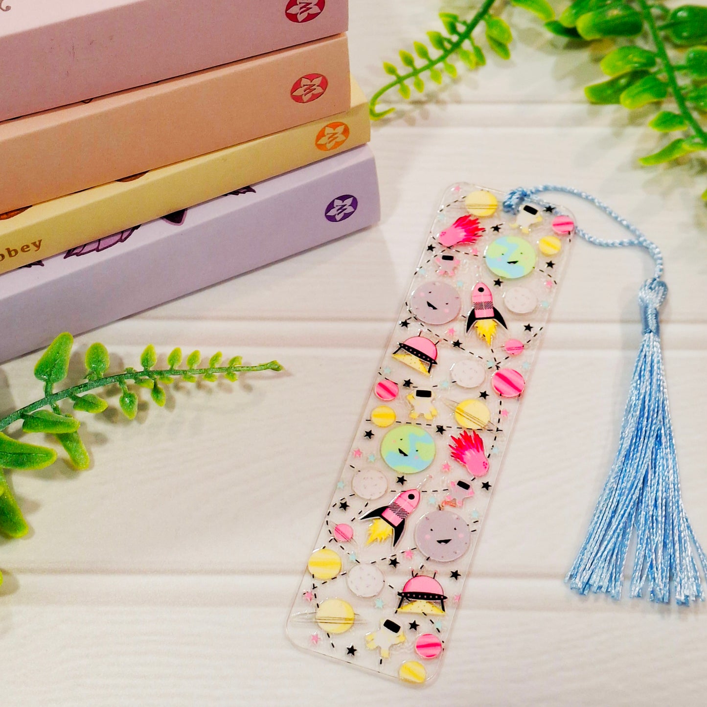 Outer space acrylic bookmark with stars and spaceships designs and a tassel, resting on a light surface.