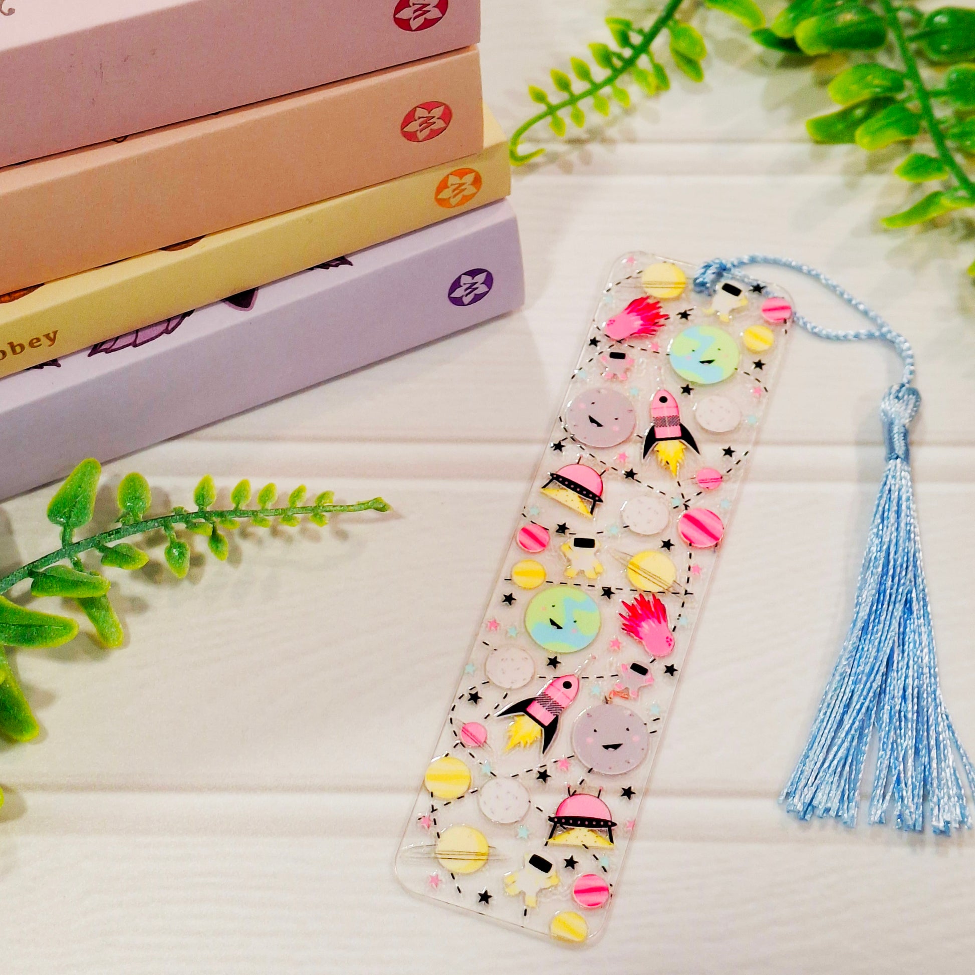 Outer space acrylic bookmark with stars and spaceships designs and a tassel, resting on a light surface.