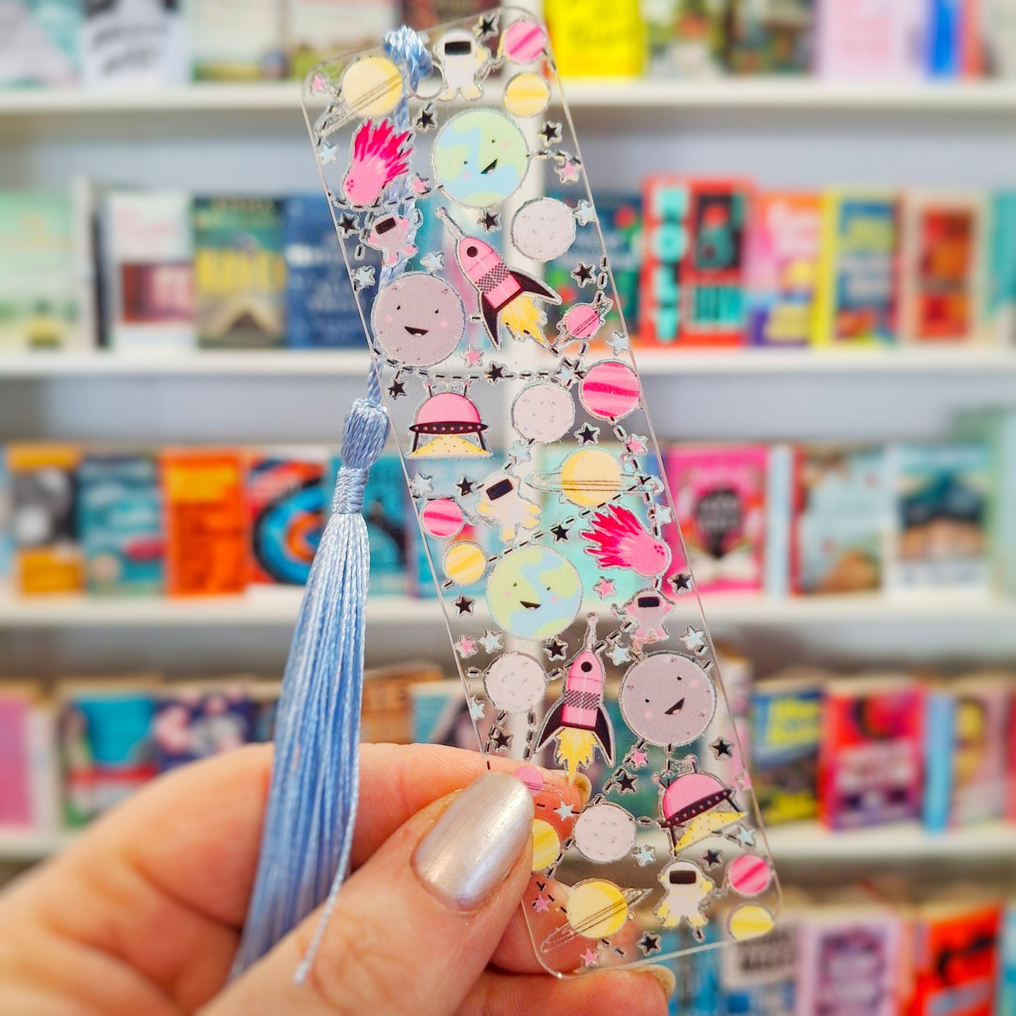 Clear acrylic bookmark featuring outer space illustrations and a tassel, held in front of a bookshelf.