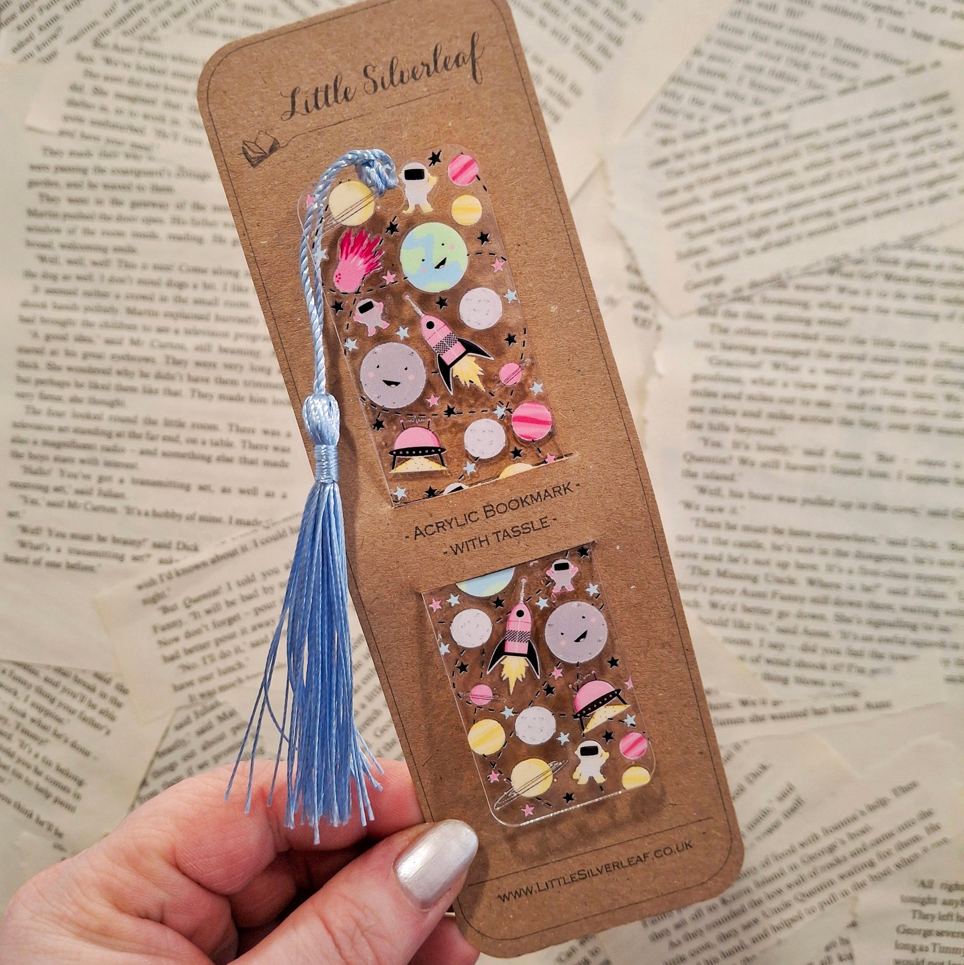 Acrylic bookmark with outer space theme and a tassel, packaged on a brown kraft backing card, held over a book page background.