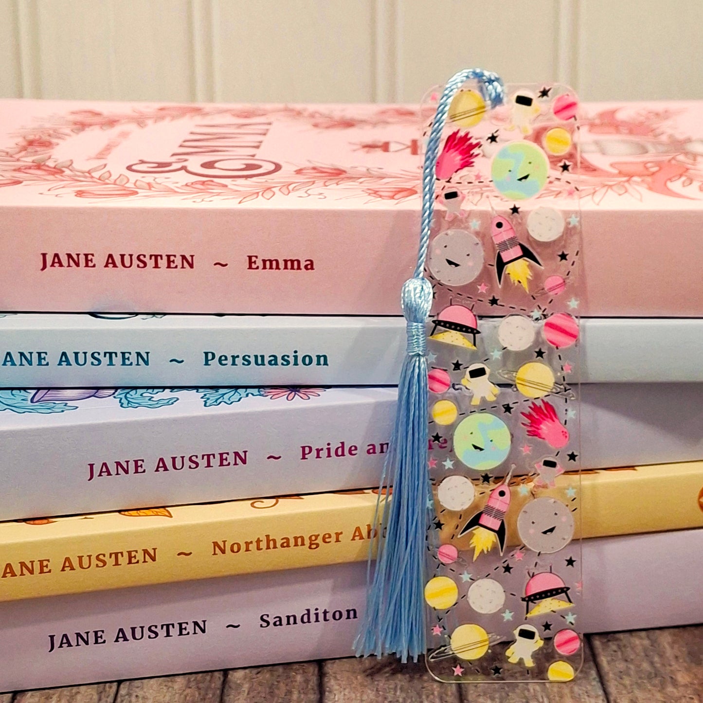 Colorful acrylic bookmark featuring outer space illustrations and a tassel, resting against a stack of rainbow coloured books.
