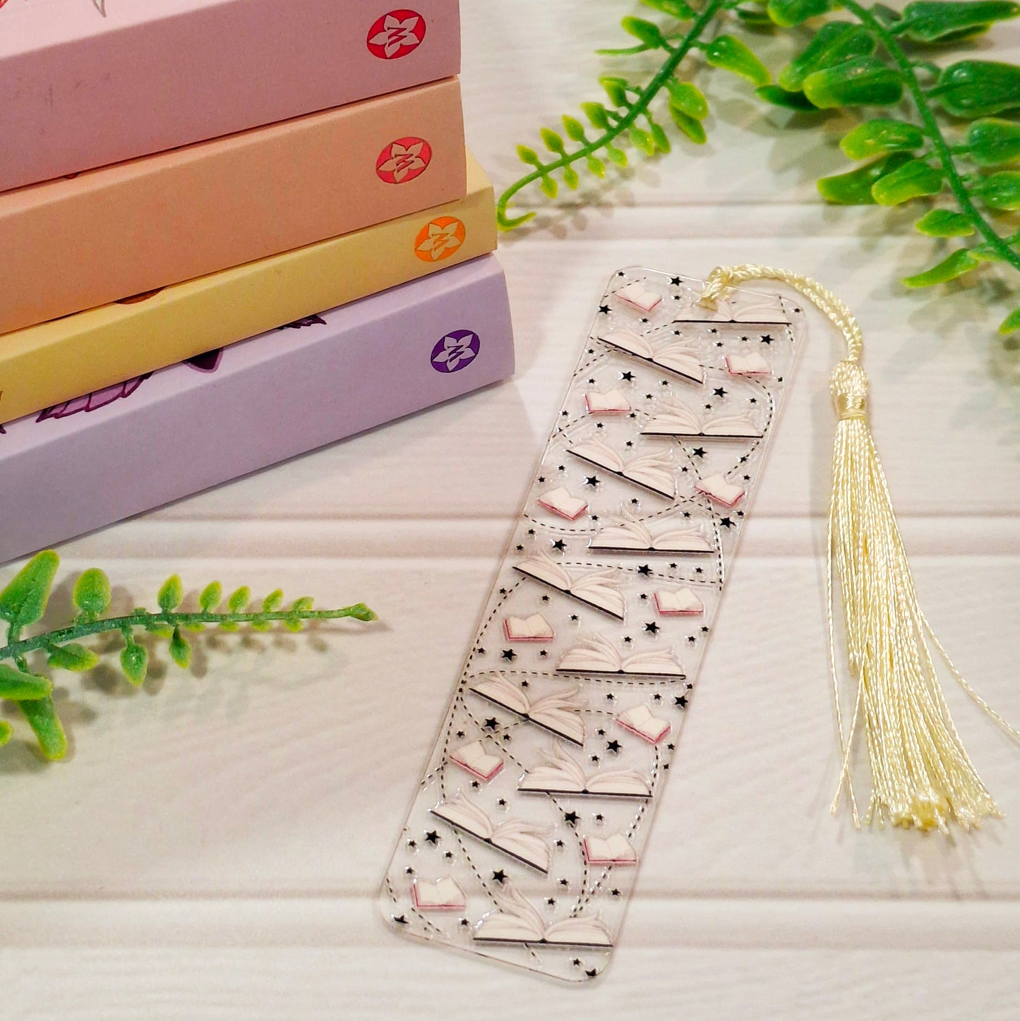 Paperback pages acrylic bookmark with tassel on a light surface, with books and green leaves.