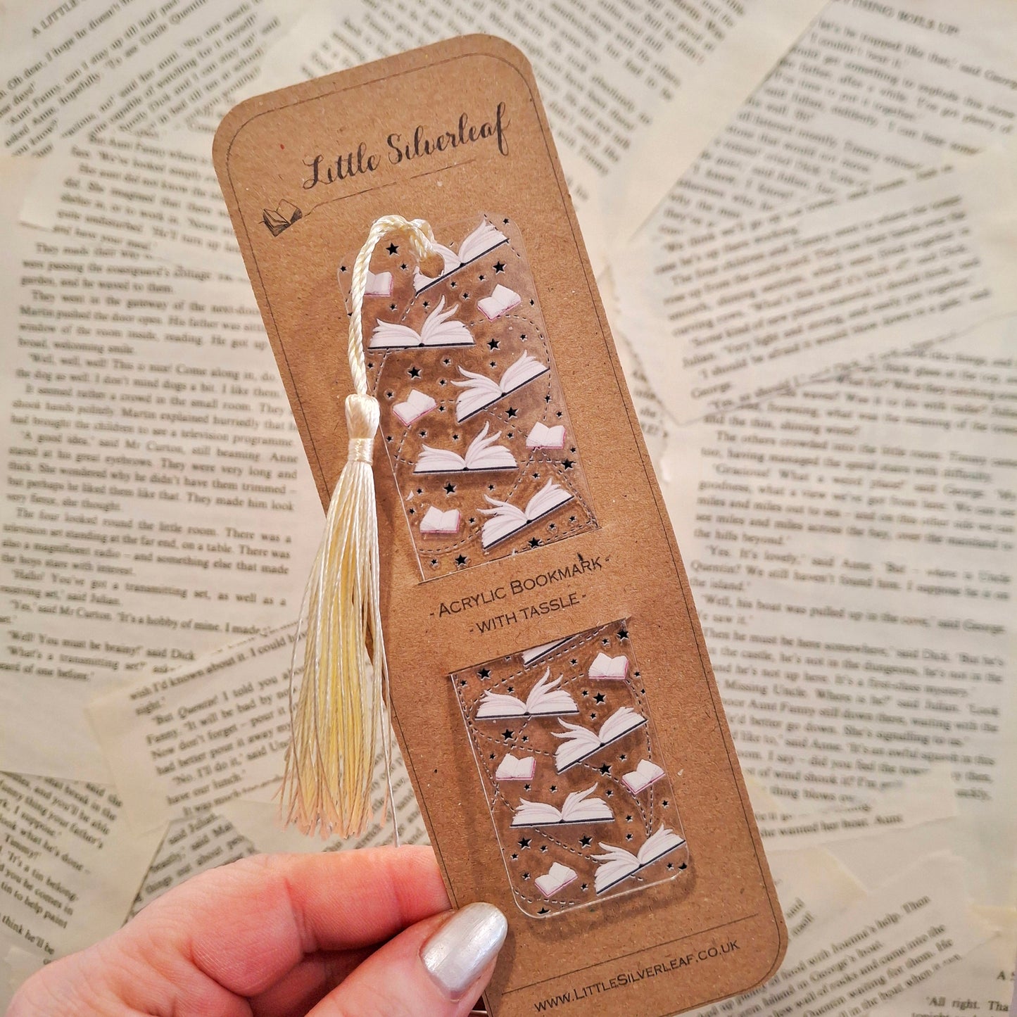 Acrylic bookmark with open book pages theme and a tassel, packaged on a brown kraft backing card, held over a book page background.