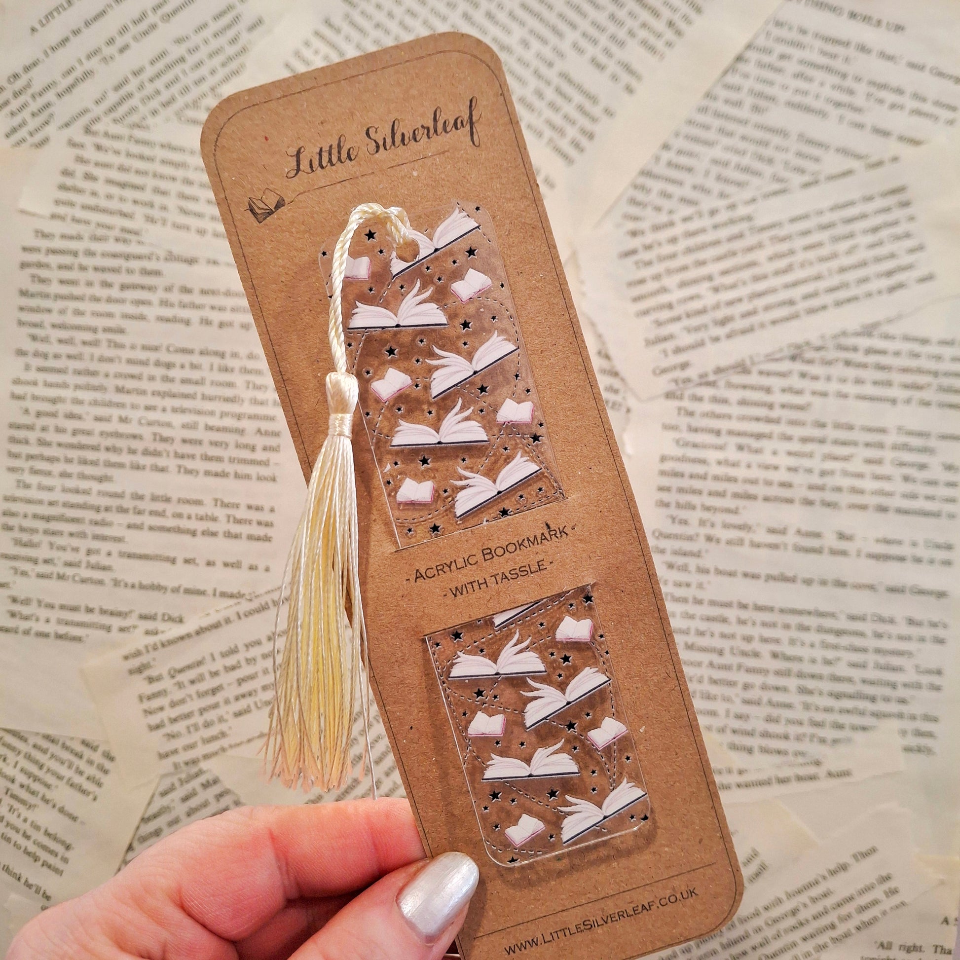 Acrylic bookmark with open book pages theme and a tassel, packaged on a brown kraft backing card, held over a book page background.