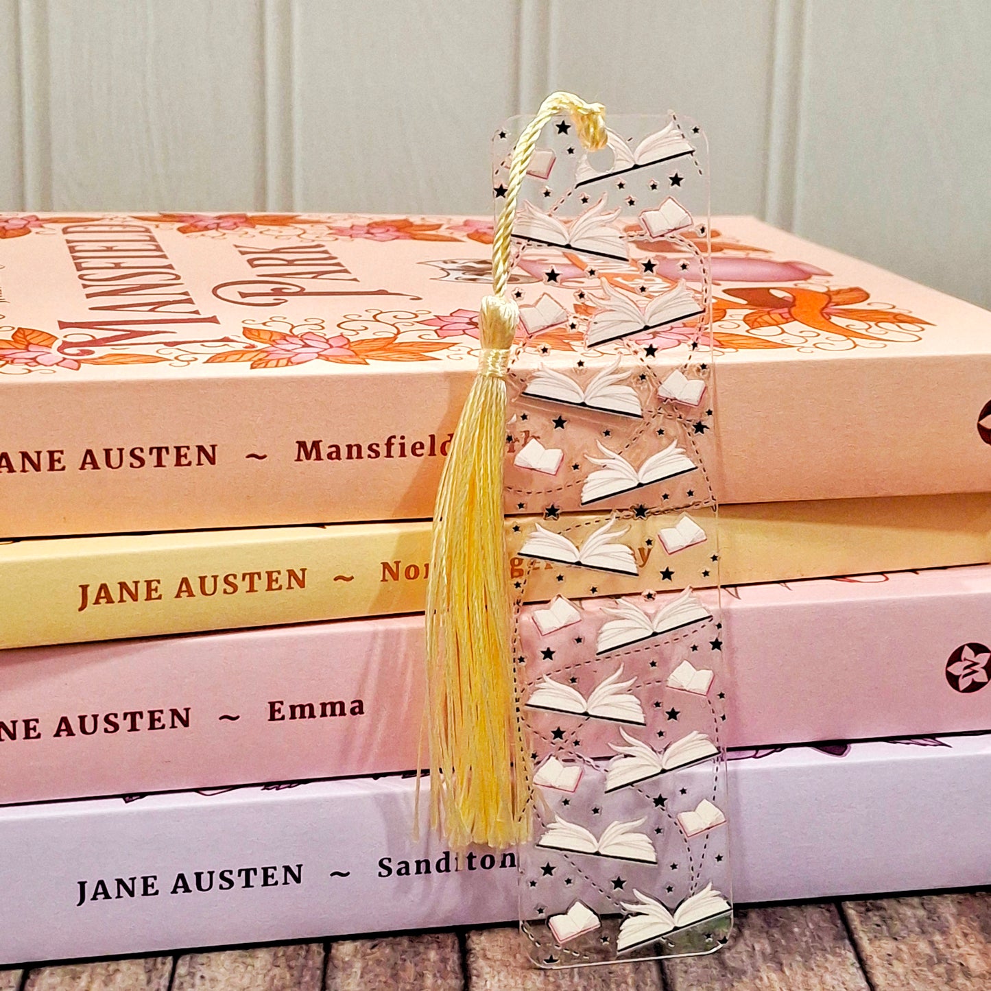 Cute bookish themed acrylic bookmark with tassel resting against a stack of books.