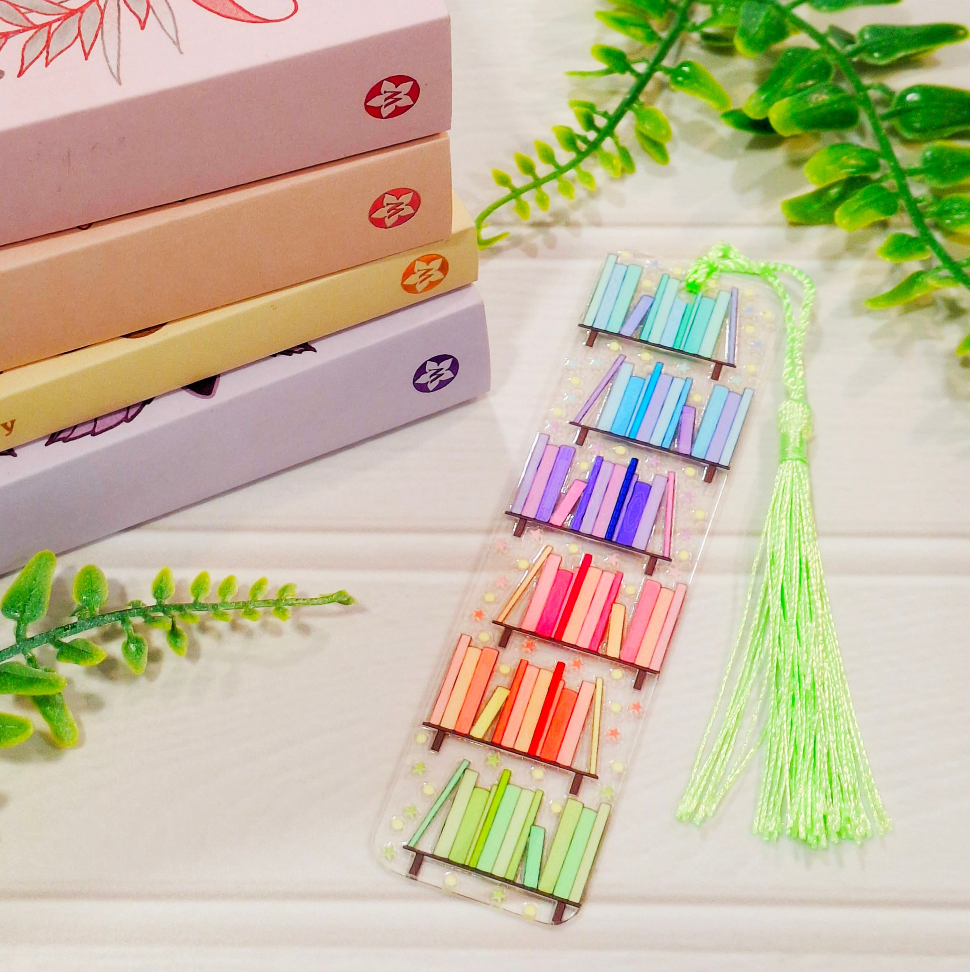 Rainbow bookshelf themed acrylic bookmark with tassel on a light surface, with books and leaves.