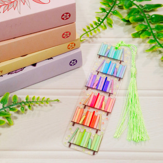 Rainbow bookshelf themed acrylic bookmark with tassel on a light surface, with books and leaves.