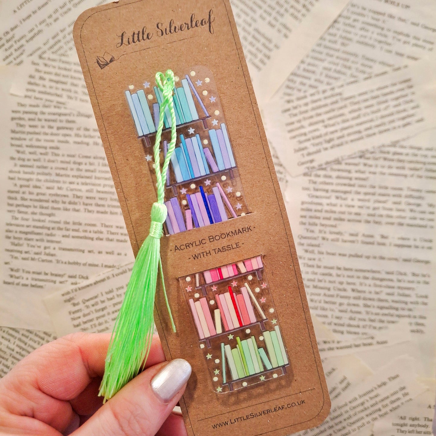 Acrylic bookmark with multicolour bookshelf design, packaged on a brown kraft backing card, held over a book page background.