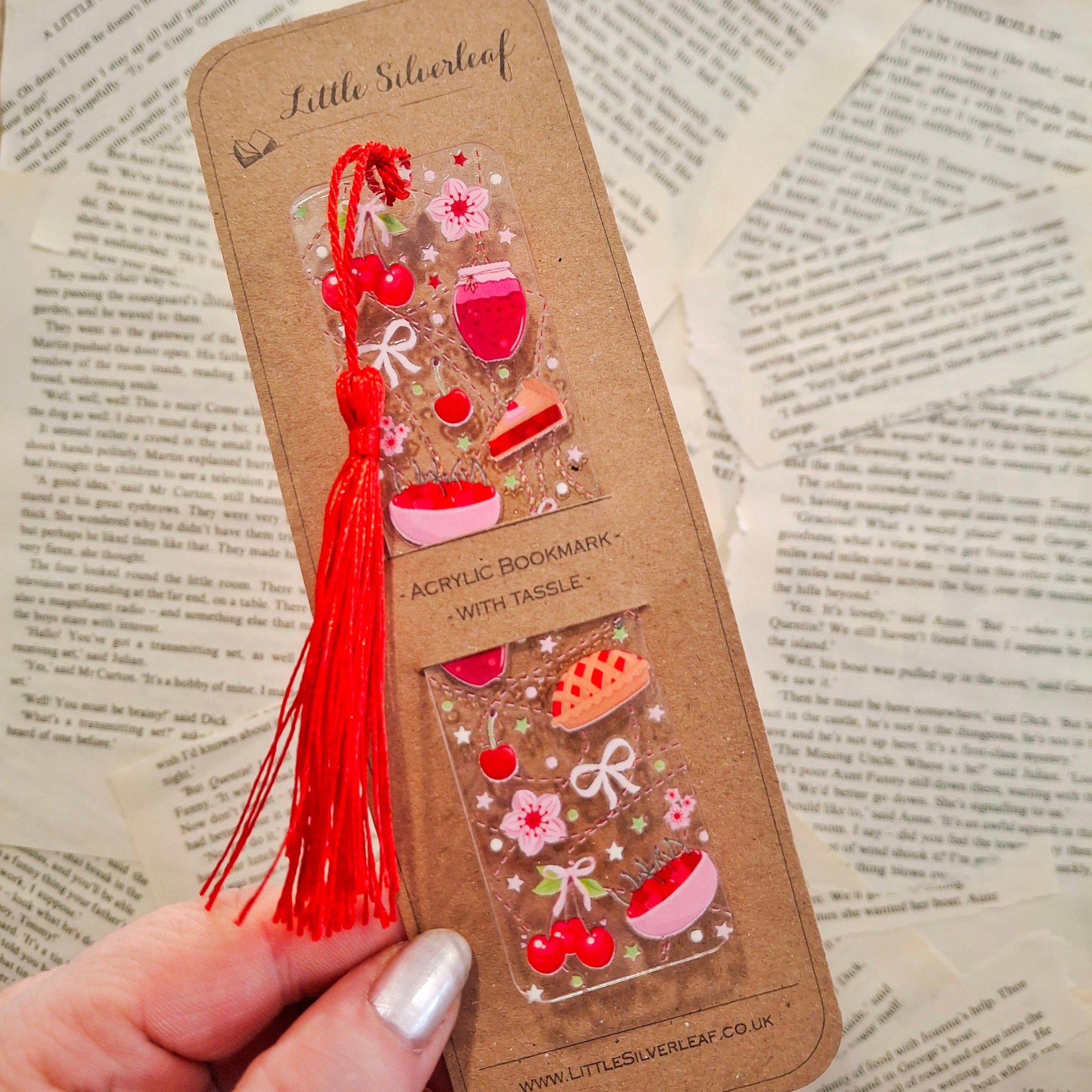 Sweet cherry pie design bookmark with tassel for cosy reading, packaged on a brown kraft backing card, held over a book page background.