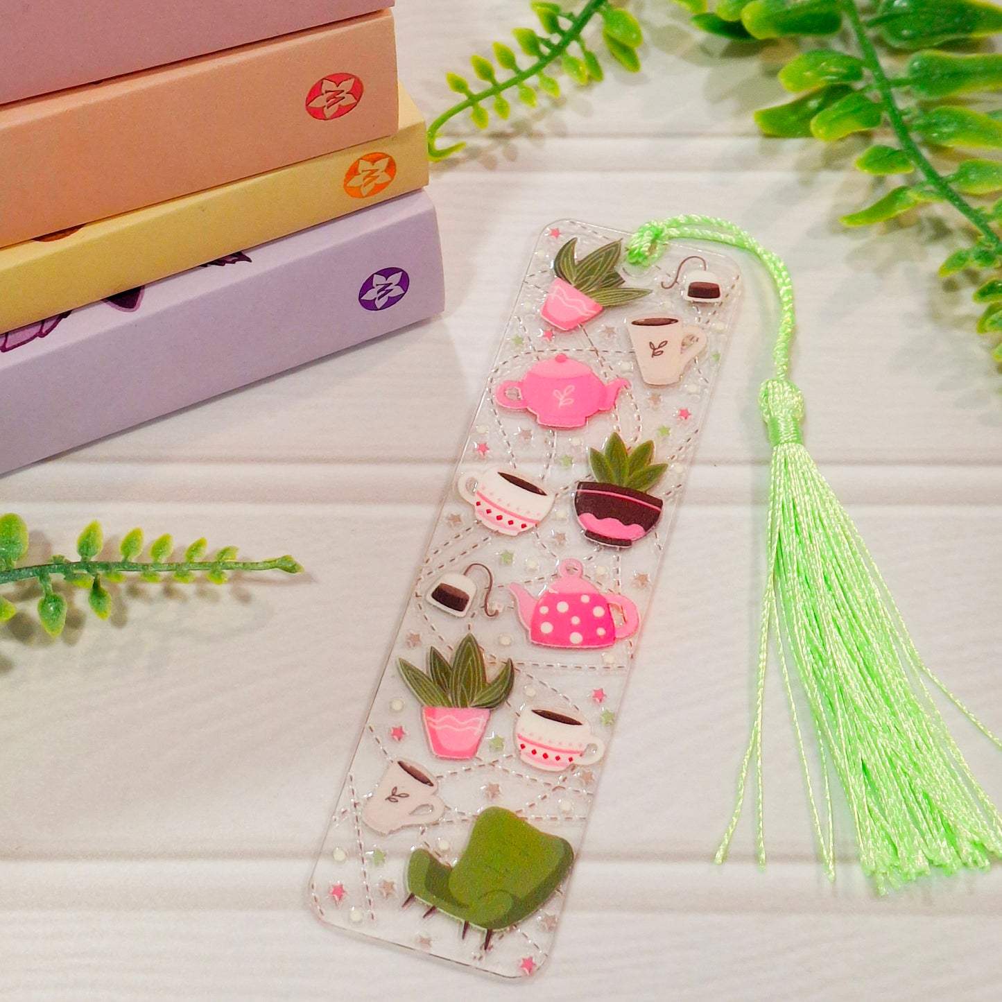 Decorative bookmark with tassel, and a houseplants, teacups and teapots design, on a light surface with books and greenery.
