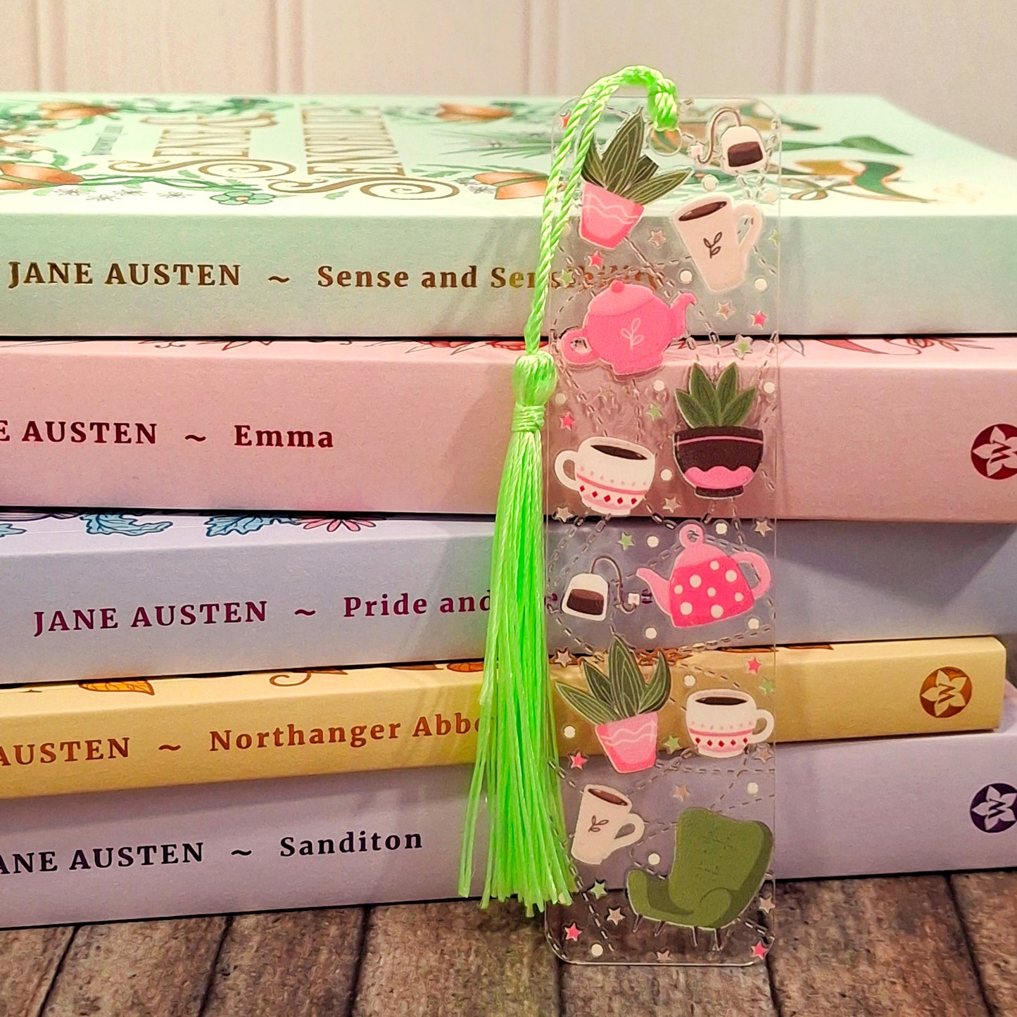 Acrylic bookmark with tassel on a stack of books, featuring illustrations of house plants, teacups and teapots.
