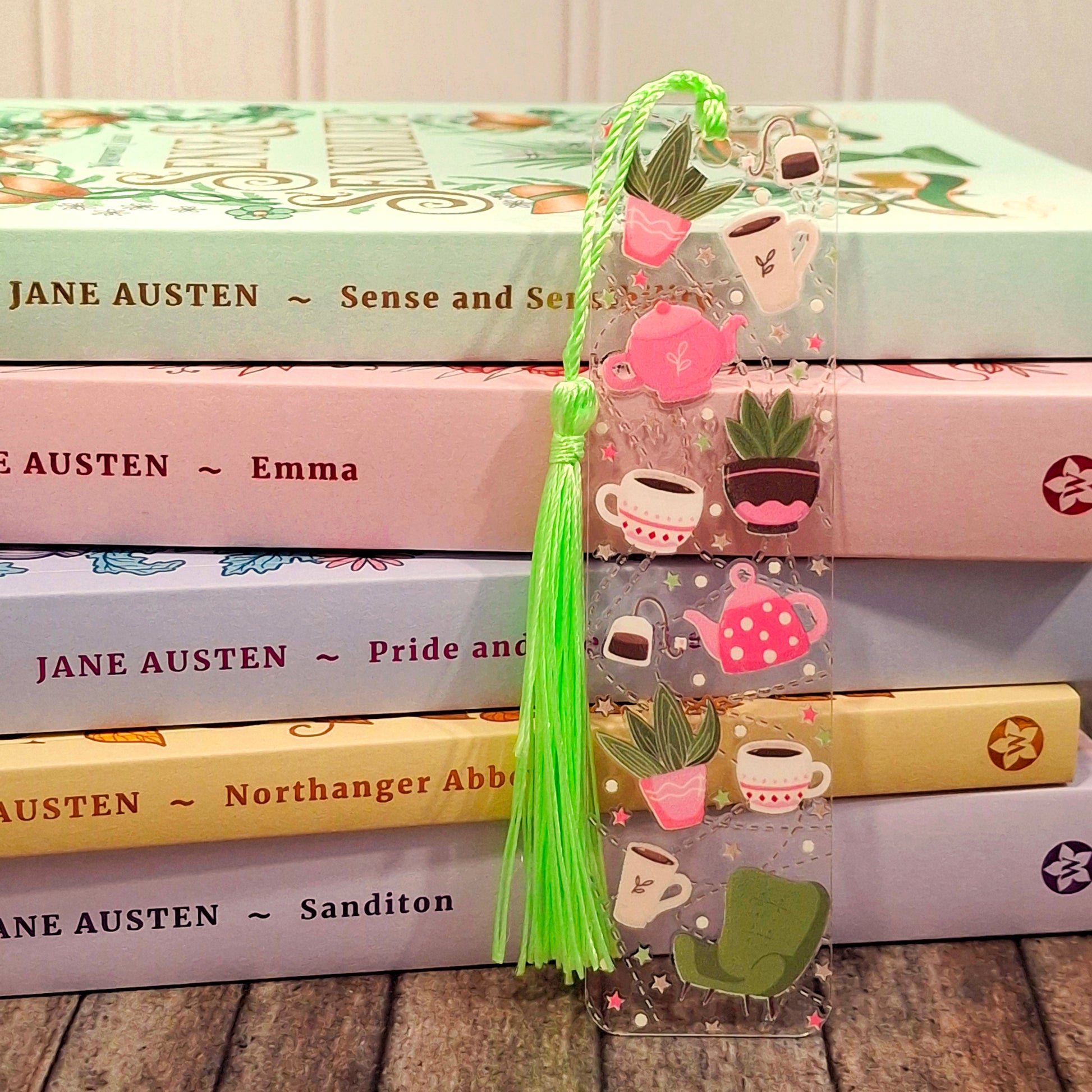 Acrylic bookmark with tassel on a stack of books, featuring illustrations of house plants, teacups and teapots.