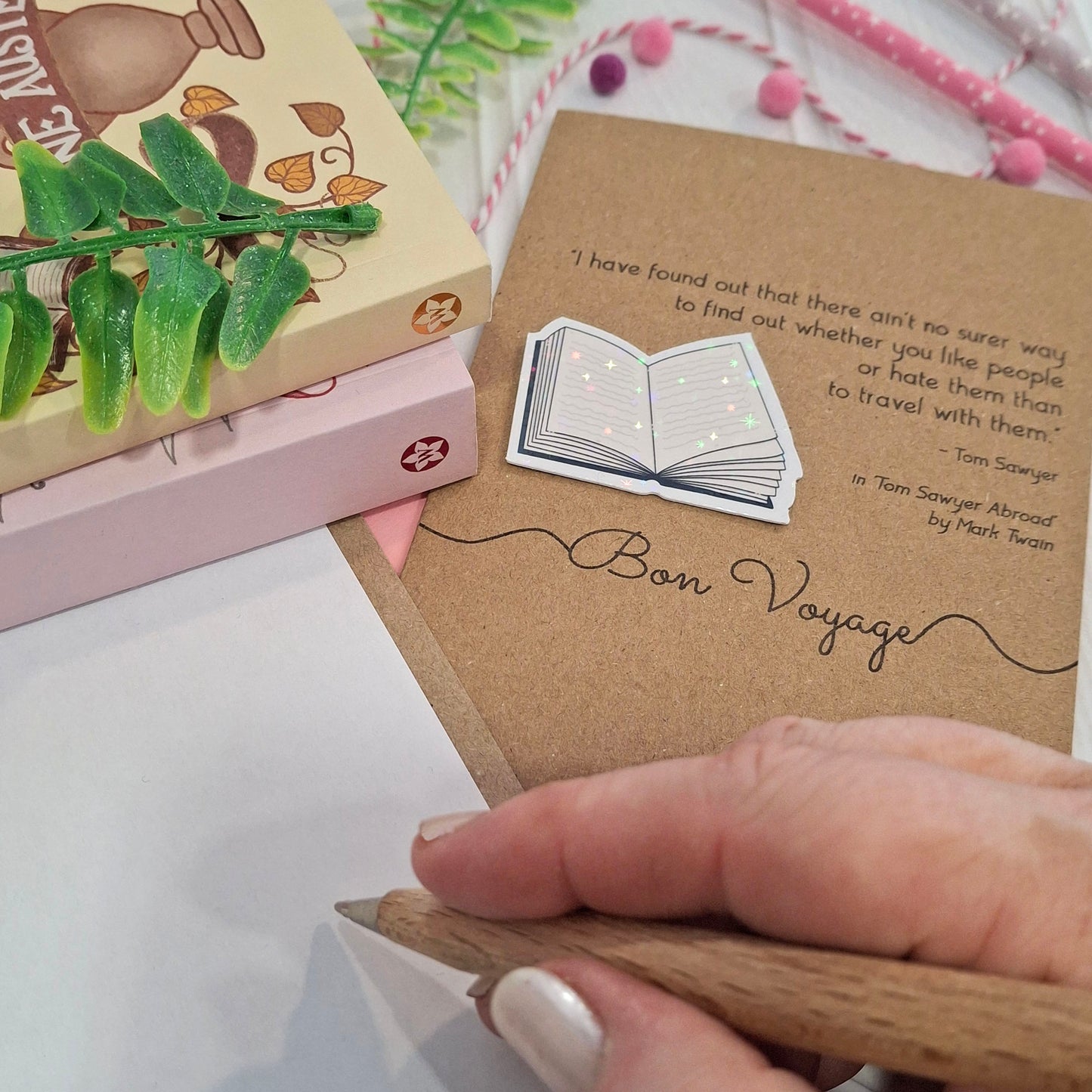 A close-up of a hand holding a wooden pen and writing inside a card. The card features a raised holographic embellishment of an open book, and reads 'Bon Voyage', with a quote from the book Tom Sawyer Abroad by mark Twain. 