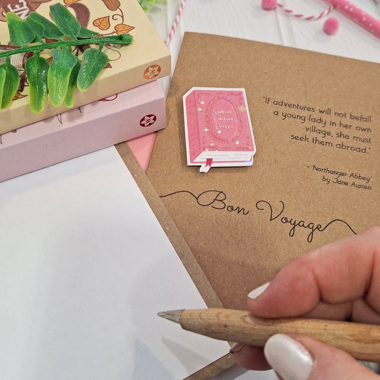 A close-up of a hand holding a wooden pencil writing inside a card. The card features a raised holographic embellishment of a pink special edition classic book, the word Bon Voyage, and a quote from Northanger Abbey by Jane Austen. The scene includes stacked books, a green leafy stem, and decorative accessories.