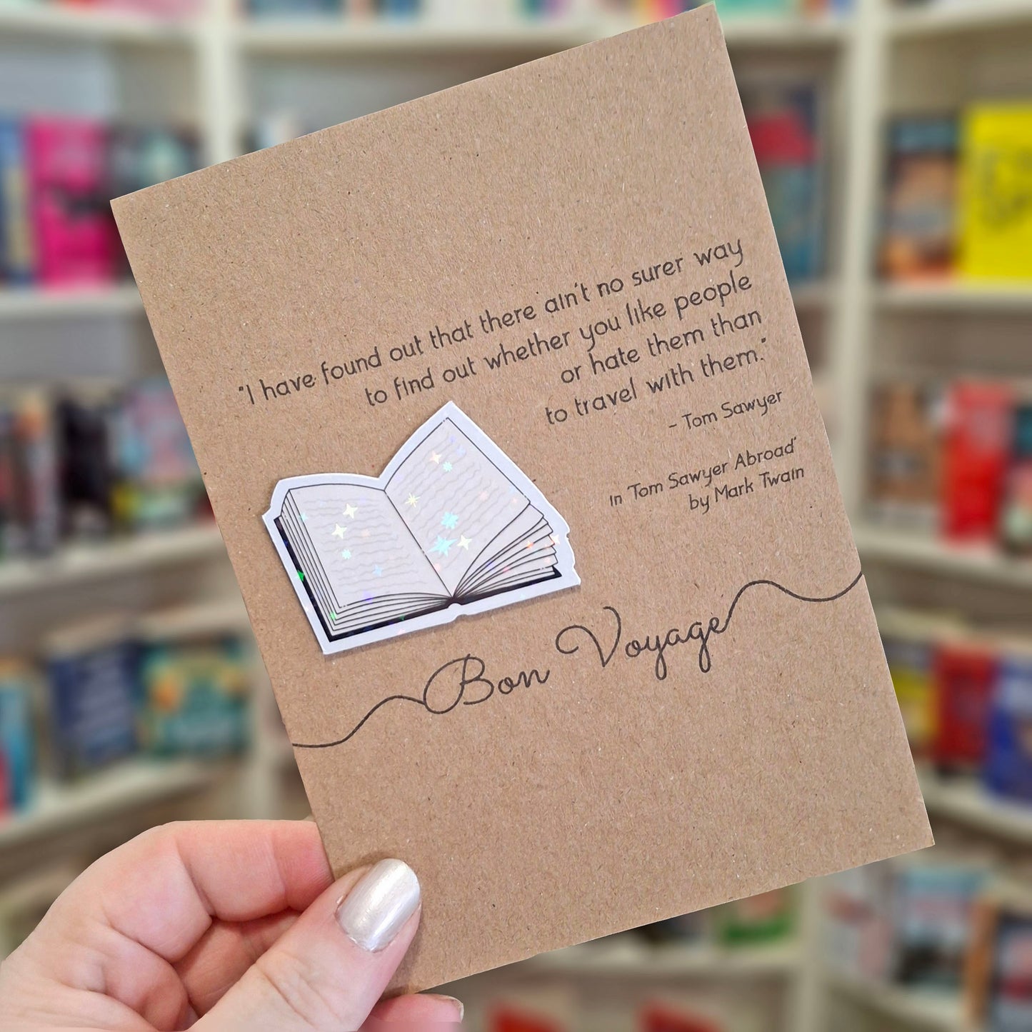 A hand holds a brown kraft bon voyage card featuring a raised holographic embellishment of an open book, and a printed quote by Mark Twain. The background shows a blurred bookshop interior.