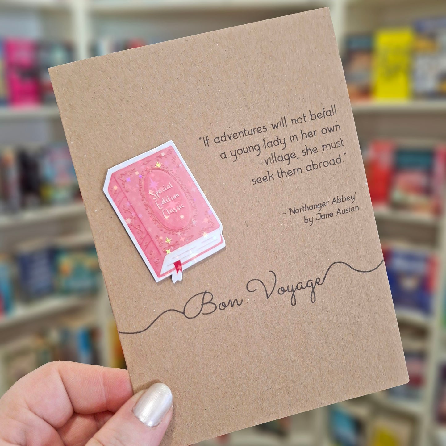 A hand holds a brown kraft bon voyage card featuring a raised holographic embellishment of a pink special edition classic book, and a printed quote by Jane Austen. The background shows a blurred bookshop interior.