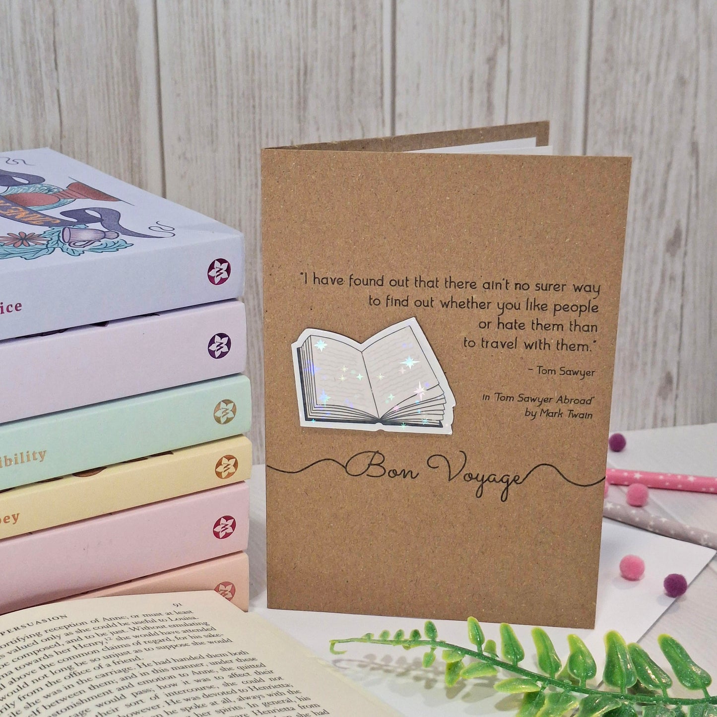 A brown kraft card styled on a tabletop with stacked pastel books, an open book, and small decorative items. The card features a raised holographic embellishment of an open book, and reads 'Bon Voyage', with a quote from the book Tom Sawyer Abroad by Mark Twain. 