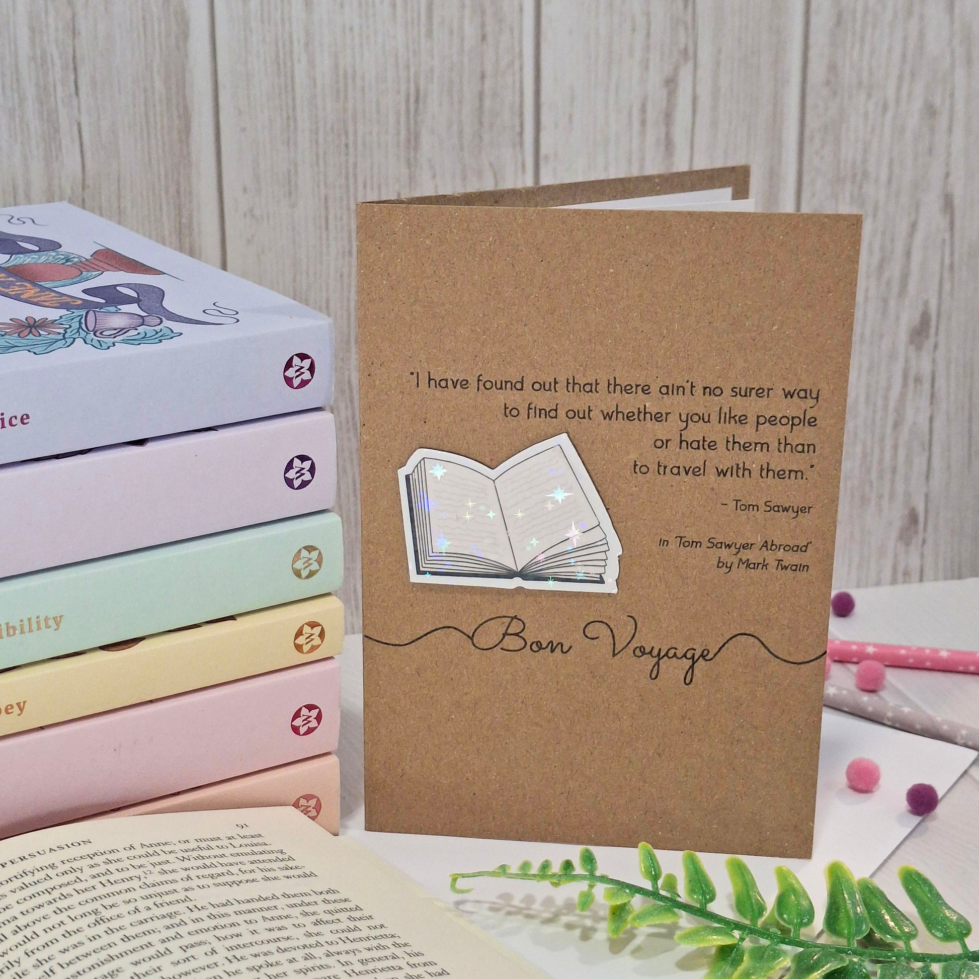 A brown kraft card styled on a tabletop with stacked pastel books, an open book, and small decorative items. The card features a raised holographic embellishment of an open book, and reads 'Bon Voyage', with a quote from the book Tom Sawyer Abroad by Mark Twain. 