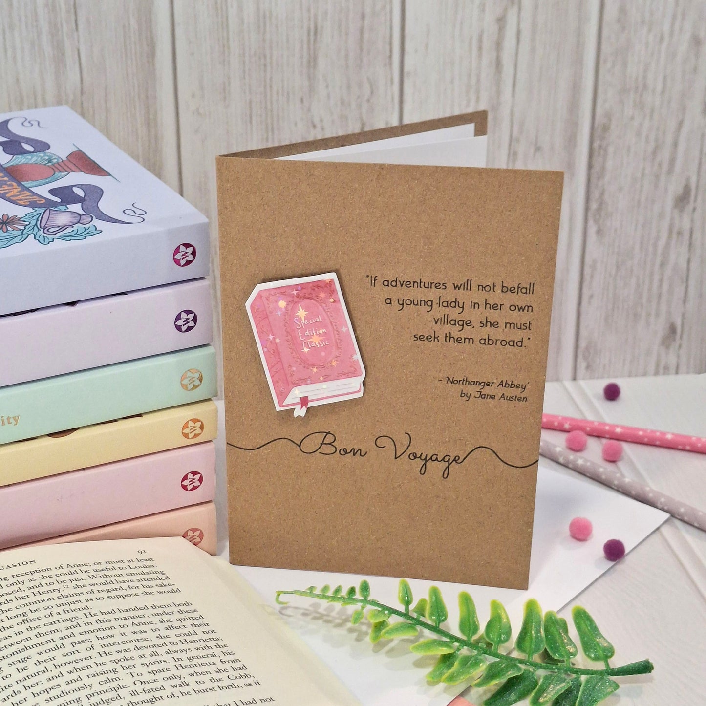A brown kraft card styled on a tabletop with books, pastel pens, and a green fern. The card features a raised holographic embellishment of a pink special edition classic book, the word Bon Voyage, and a quote from Northanger Abbey by Jane Austen.