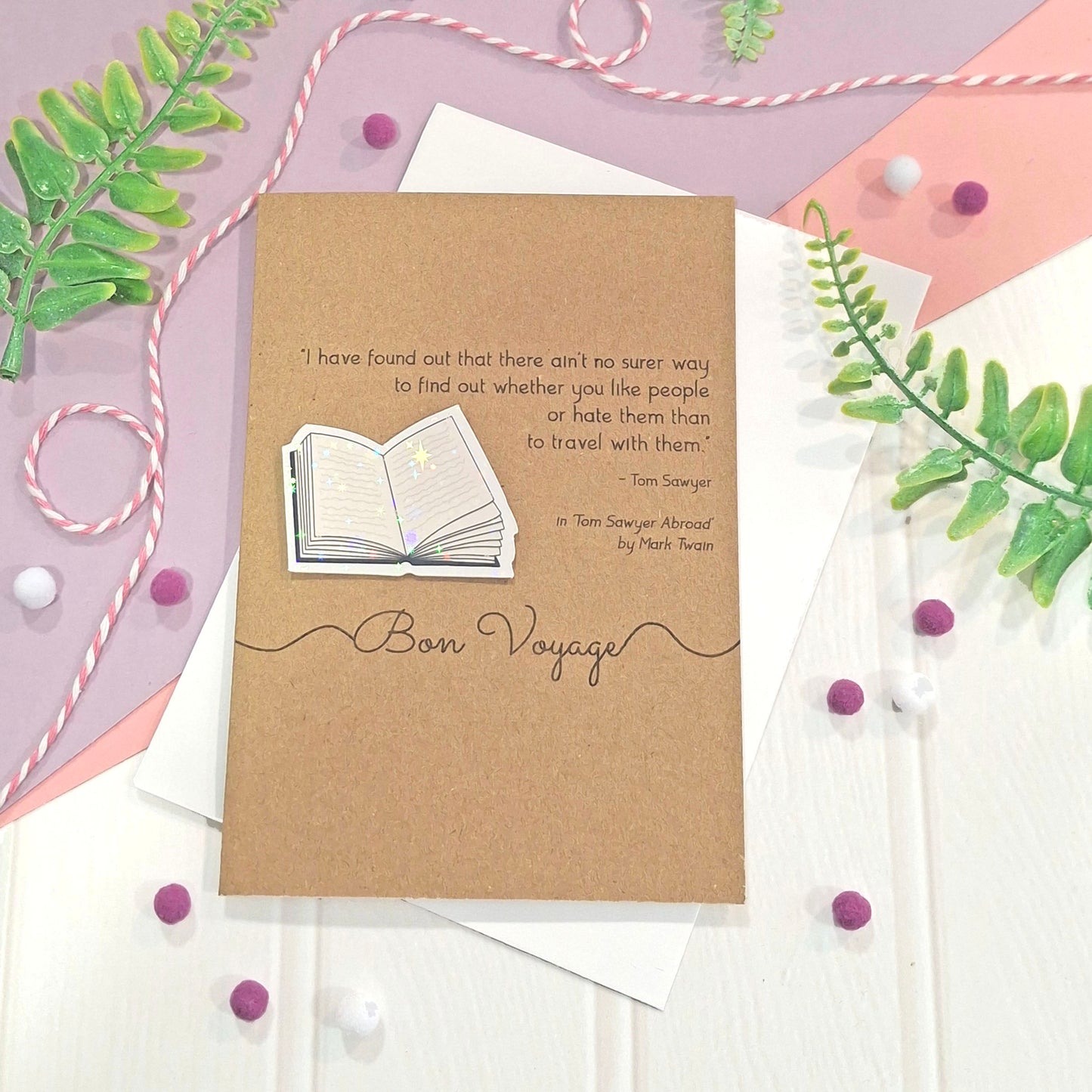 A kraft card with a travel-themed quote from the book Tom Sawyer by Mark Twain and the words 'Bon Voyage' on it, topped with a raised, holographic, illustrated open book embellishment and styled in a flat lay with white envelopes on a pastel background.
