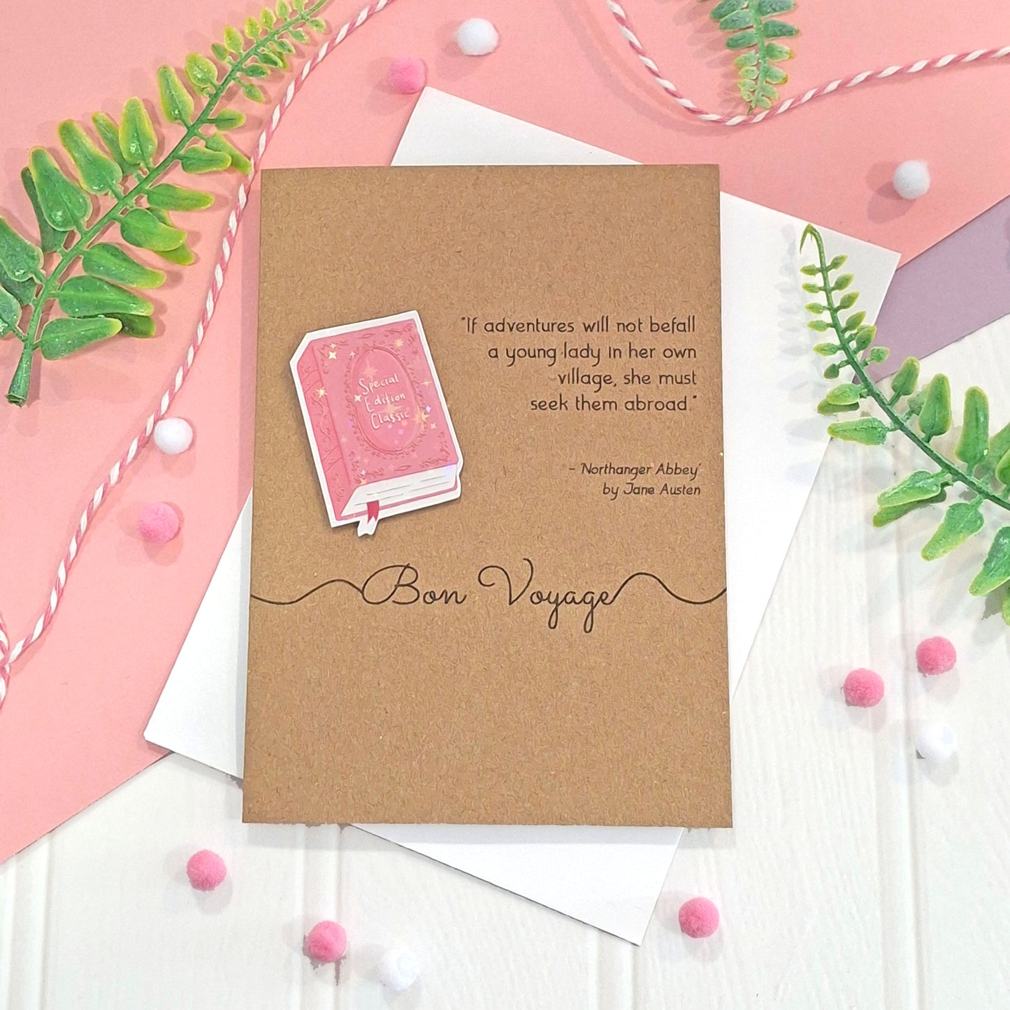 A kraft card with a quote from Northanger Abbey by Jane Austen, and the words 'Bon Voyage'. The card features a raised, holographic, illustrated story book embellishment in pink, and is styled on a white and pink background with green leaves and pink pom-poms.