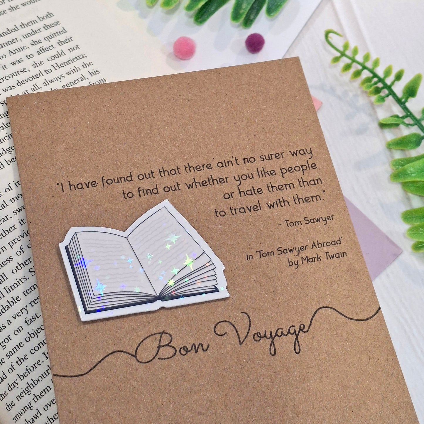 A kraft card styled on top of an open book with decorative greenery and pom-poms. The card features a raised holographic embellishment of an open book, and reads 'Bon Voyage', with a quote from the book Tom Sawyer Abroad by Mark Twain. 