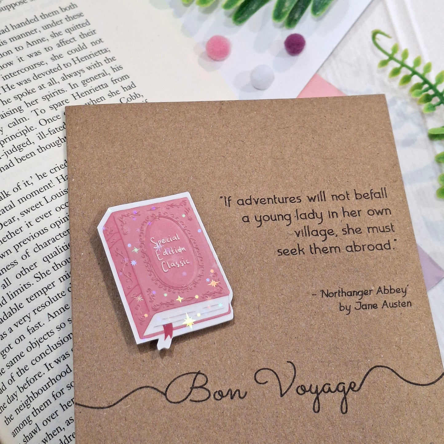 A brown kraft card styled on top of an open book with green leaves and pom-poms in the background. The card features a raised holographic embellishment of a pink special edition classic book, the word Bon Voyage, and a quote from Northanger Abbey by Jane Austen.