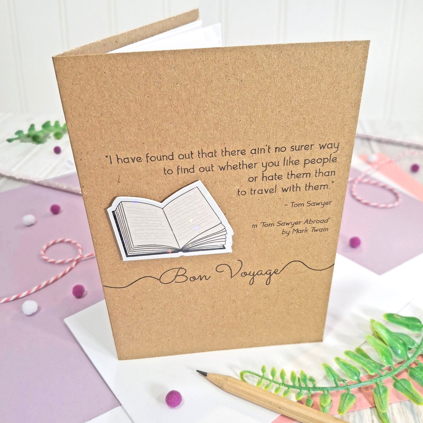 A handmade kraft card with a printed quote from the book Tom Sawyer by Mark Twain, and the words 'Bon Voyage'. The card features a raised, holographic, illustrated open book embellishment, and is shown standing upright.