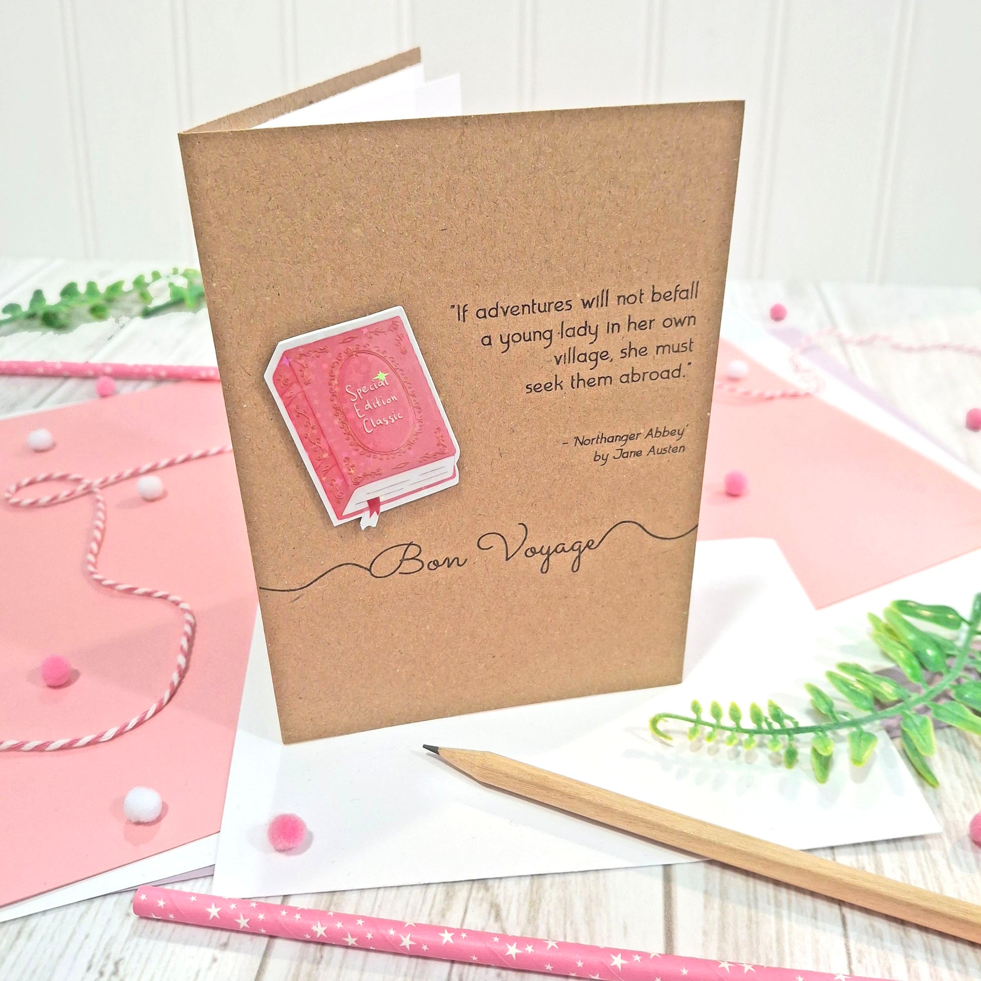 A handmade greeting card in brown kraft card with a raised, holographic, illustrated story book embellishment in pink, the words 'Bon Voyage', and a quote from Northanger Abbey by Jane Austen, displayed on a styled tabletop with pink paper, pens, and greenery.