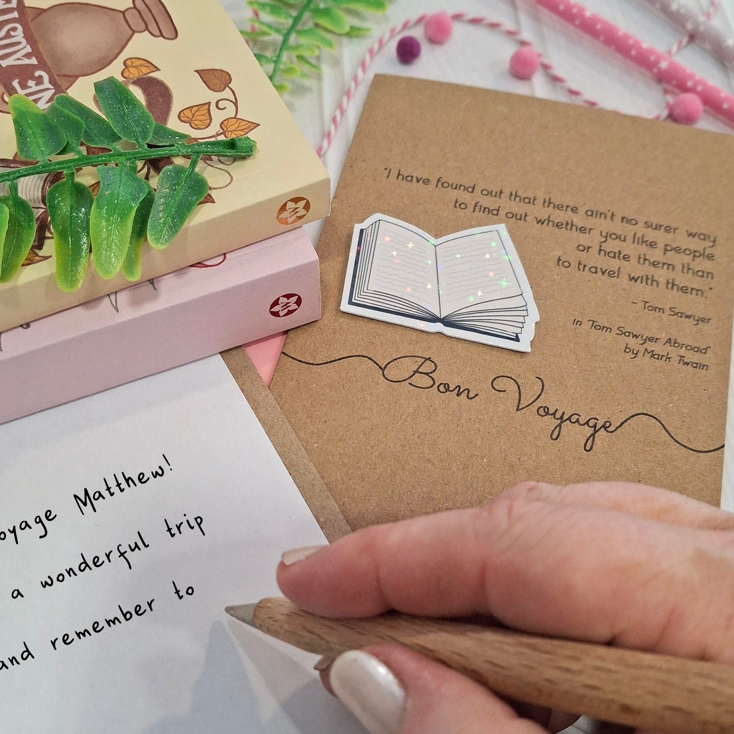 A close-up of a hand holding a wooden pen and writing a goodbye message inside a card. The card features a raised holographic embellishment of an open book, and reads 'Bon Voyage', with a quote from the book Tom Sawyer Abroad by Mark Twain. 