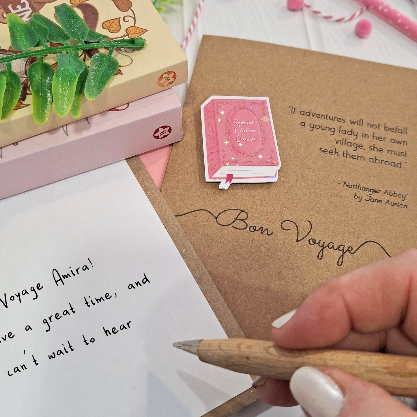 A close-up of a hand holding a wooden pencil writing a goodbye message inside a card. The card features a raised holographic embellishment of a pink special edition classic book, the word Bon Voyage, and a quote from Northanger Abbey by Jane Austen.. The scene includes stacked books, a green leafy stem, and decorative accessories.