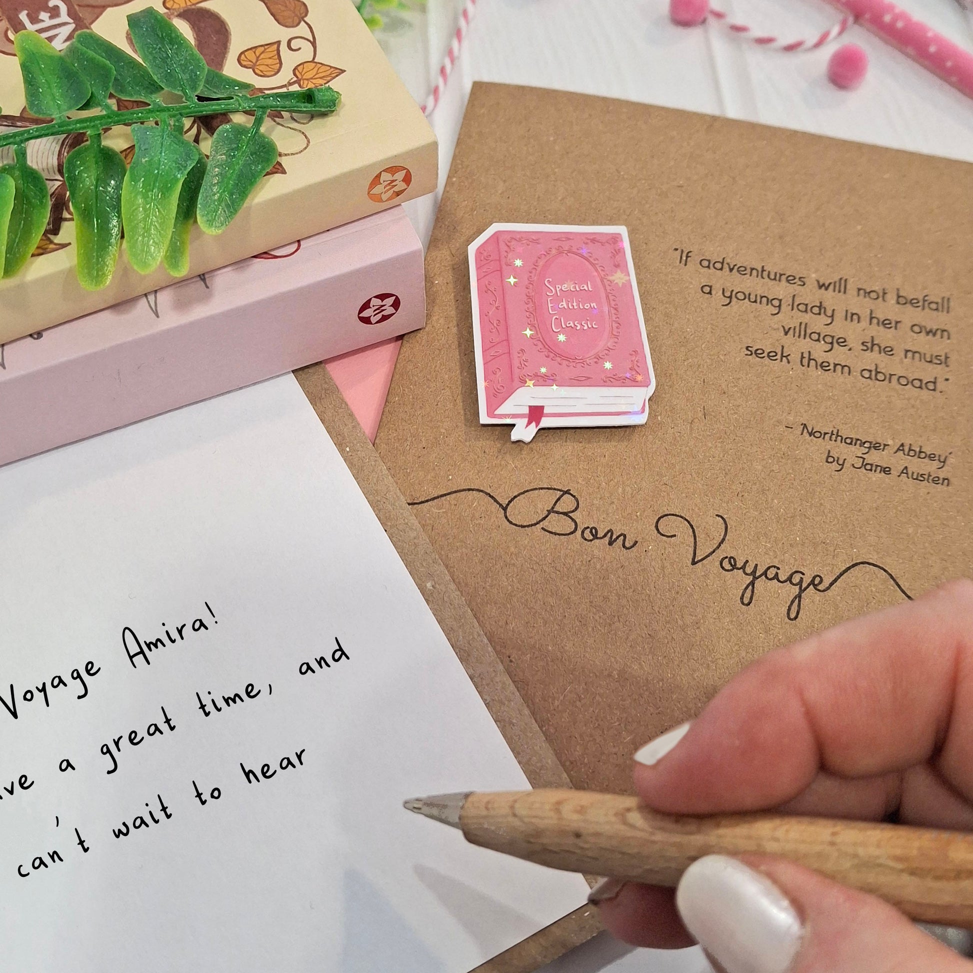 A close-up of a hand holding a wooden pencil writing a goodbye message inside a card. The card features a raised holographic embellishment of a pink special edition classic book, the word Bon Voyage, and a quote from Northanger Abbey by Jane Austen.. The scene includes stacked books, a green leafy stem, and decorative accessories.