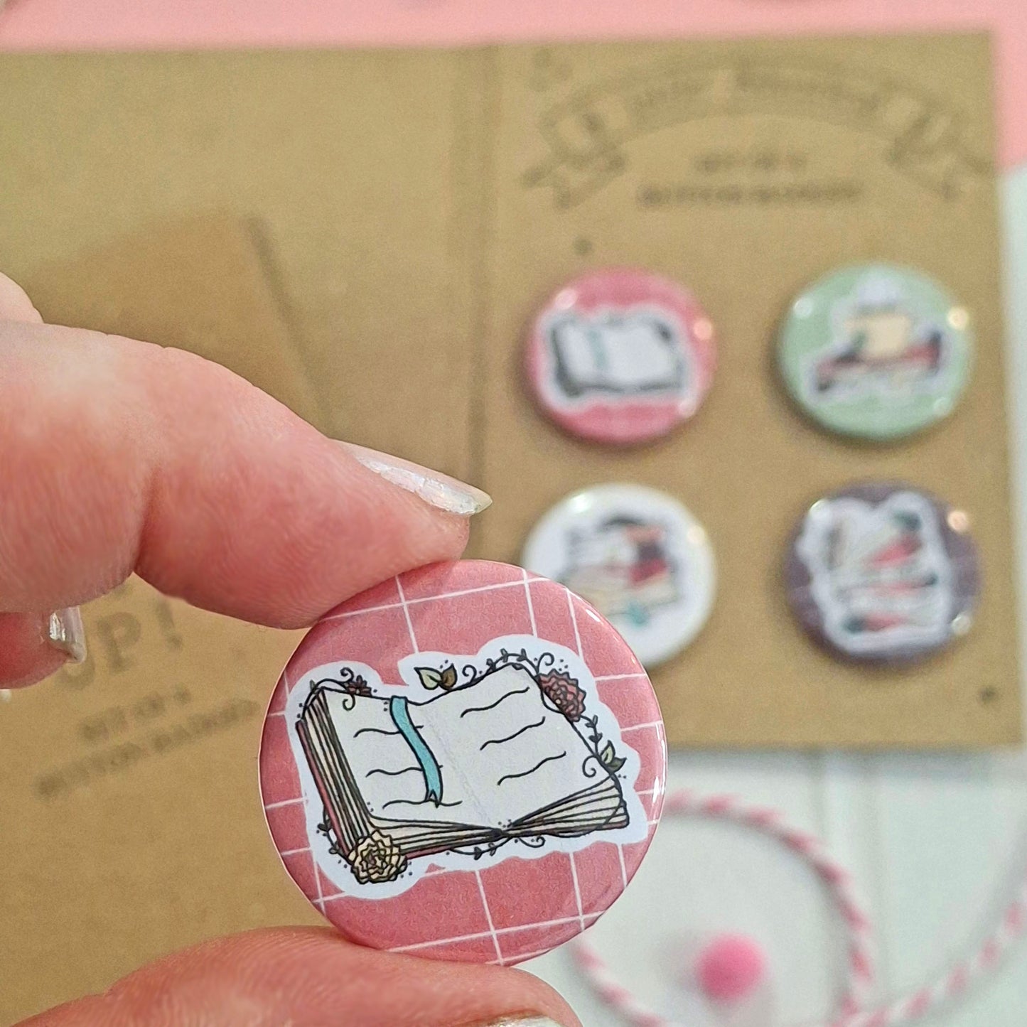 A hand holds a round pinback button featuring an illustrated open book with a blue ribbon on a pink grid background. Other similar buttons are displayed on a kraft card in the blurred background.