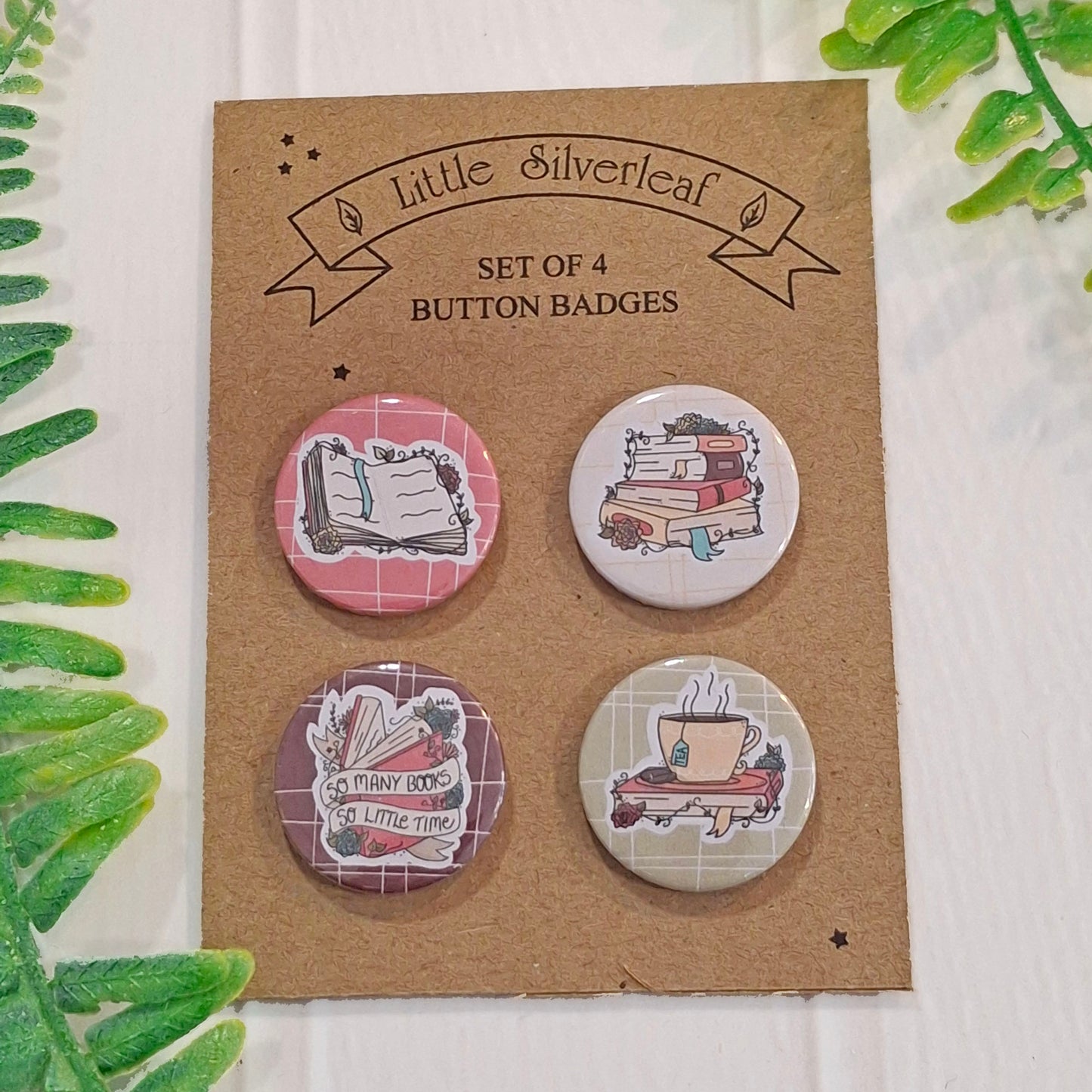 A set of four round button badges displayed on a brown kraft card labeled 'Little Silverleaf Set of 4 Button Badges.' The badges feature illustrated book-themed designs, including an open book, stacked books, a book with the phrase 'So many books so little time,' and a tea cup.