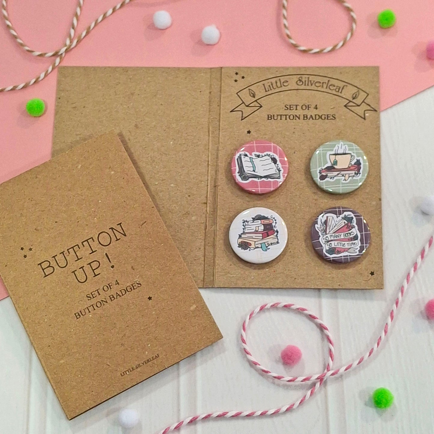 A set of four round button badges displayed on kraft paper backing cards, with the cards arranged on a pink and white surface with decorative twine and pom-poms. The badges feature illustrated book-themed designs, including a book, a tea cup, stacked books, and a book with a ribbon reading 'so many books, so little time'.