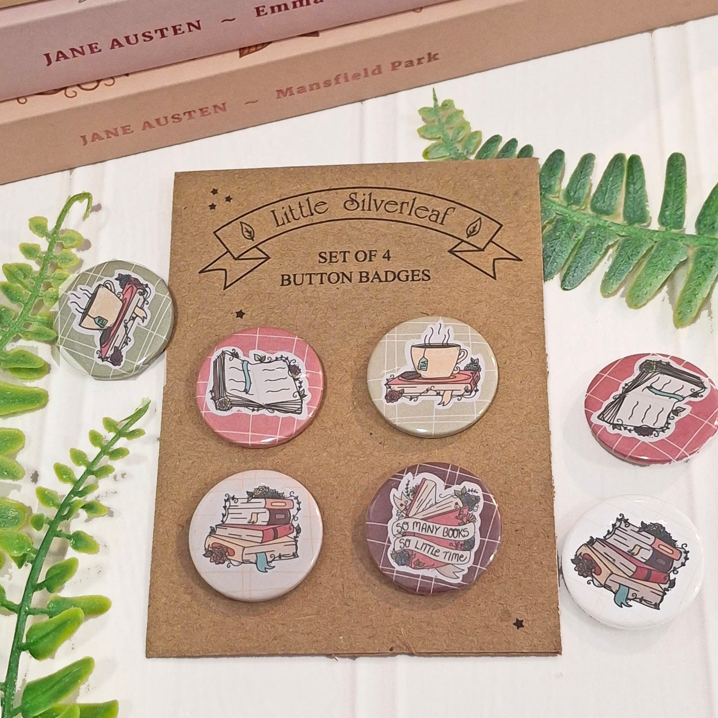 A set of four round button badges displayed on a brown kraft card labelled 'little Silverleaf SET OF 4 BUTTON BADGES.' The badges feature illustrated book-themed designs, including open books, tea cups, and stacked books.