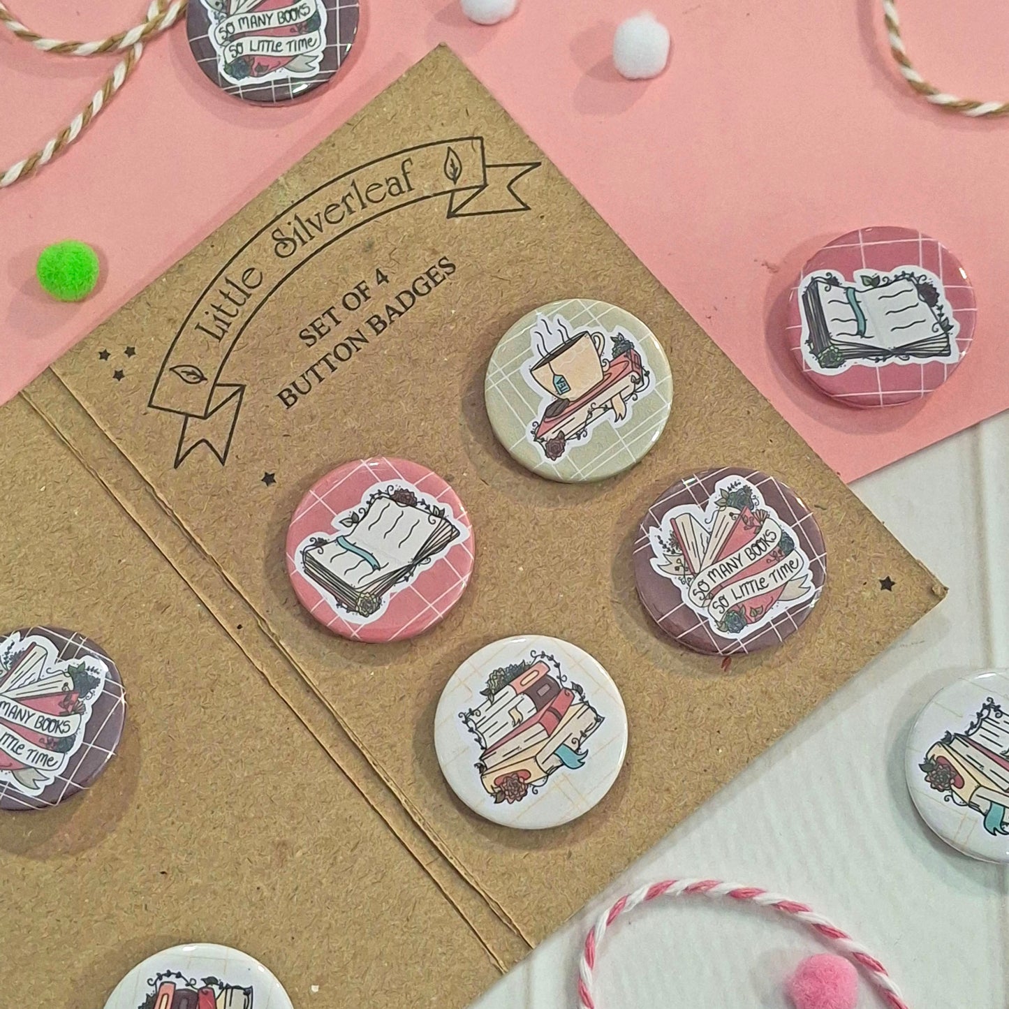 A set of four round button badges displayed on a kraft card labeled 'Little Silverleaf Set of 4 Button Badges.' The badges feature illustrated book covers on a pink, white, and brown background with decorative pom-poms and twine.