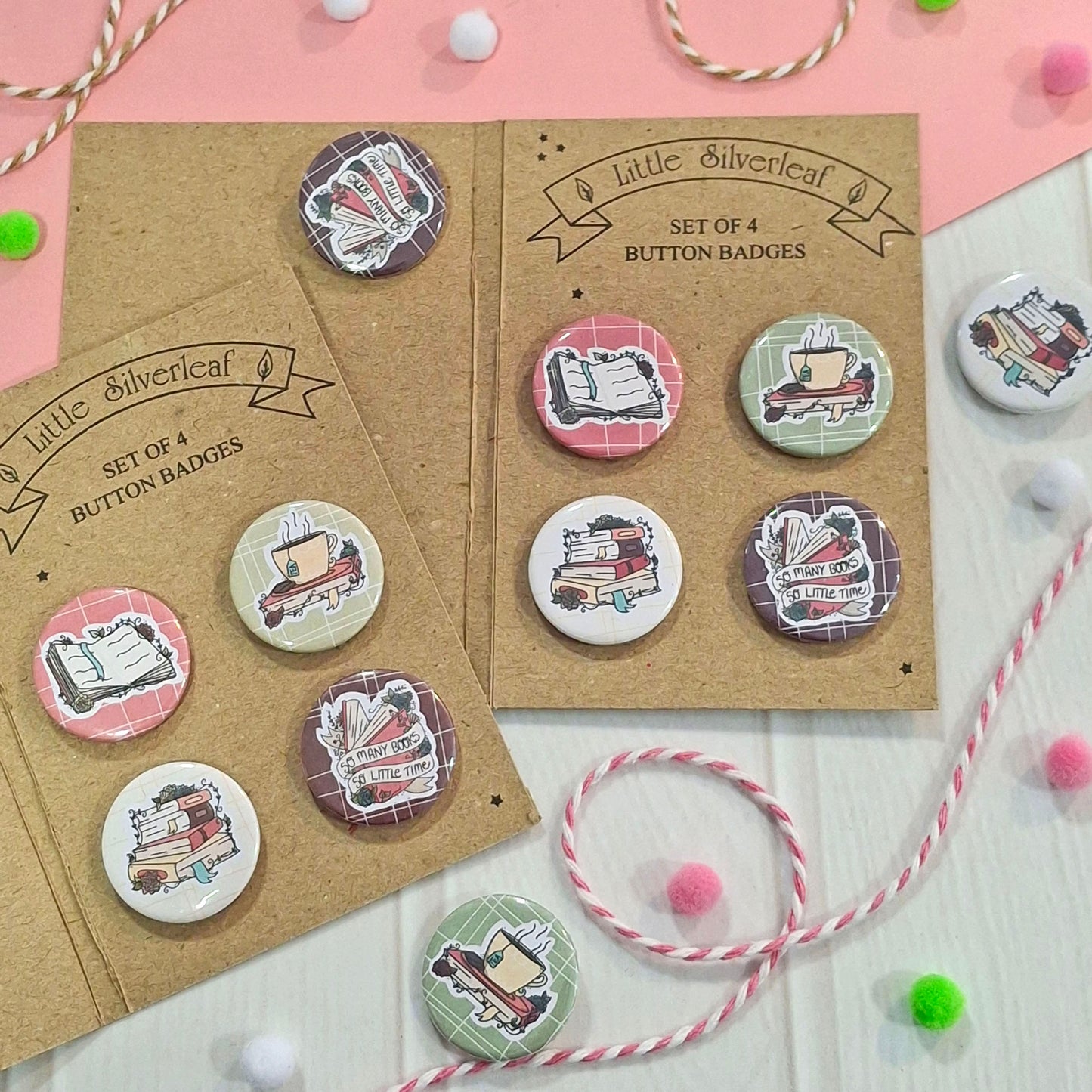Two kraft display cards holding eight round button badges with illustrated book designs, arranged on a pink and white surface with decorative string and pom-poms.