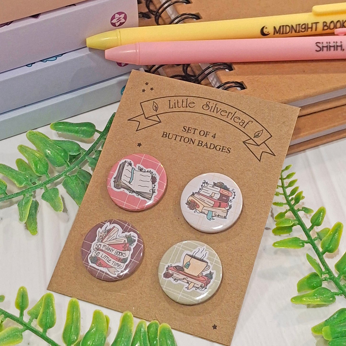 A set of four illustrated button badges displayed on a brown kraft card labelled 'Little Silverleaf' and 'Set of 4 Button Badges.' The badges feature book-themed designs in pink, white, brown, and beige tones.